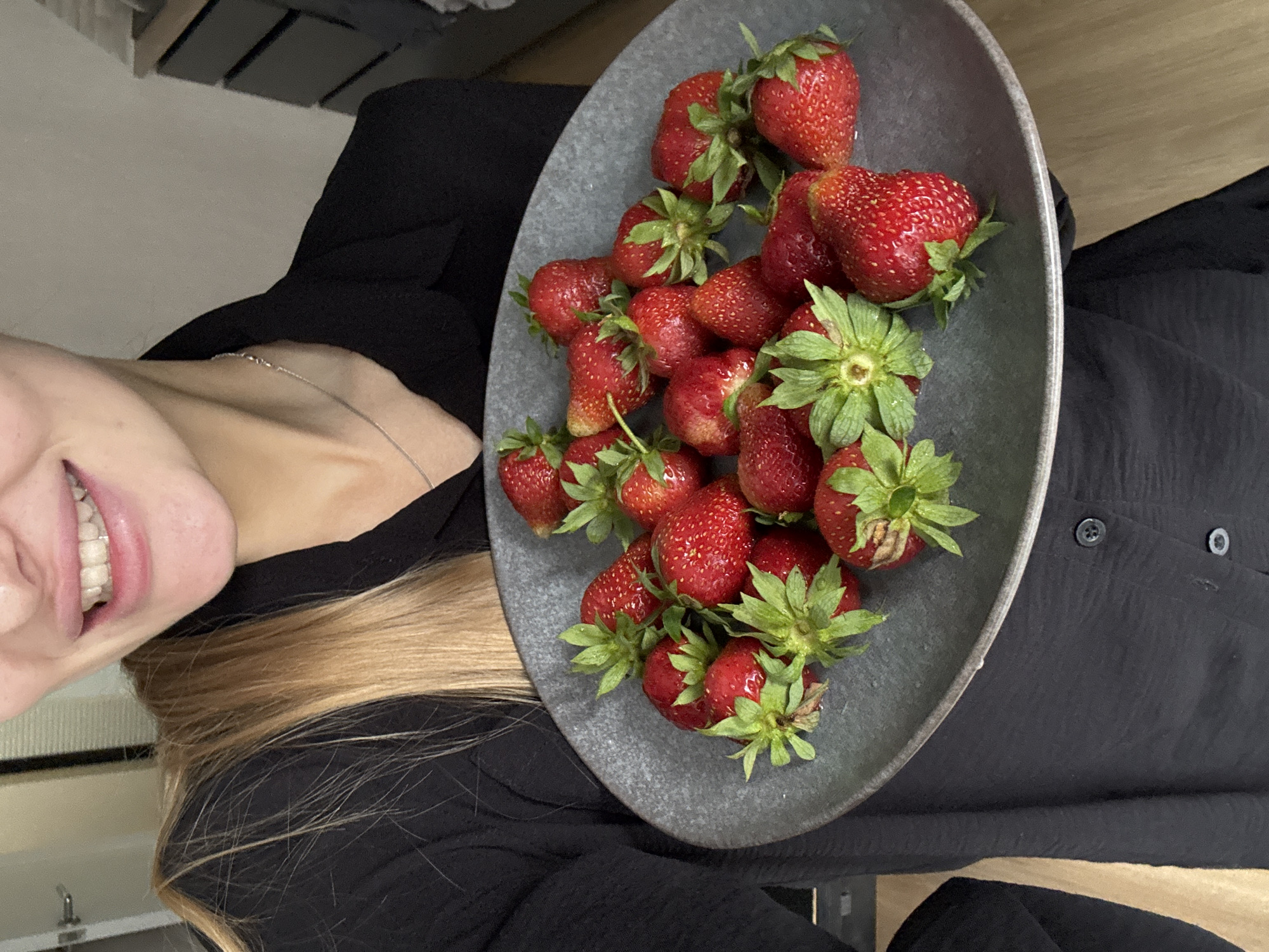 In a close-up shot, a person holding a dark gray plate filled with fresh strawberries is visible. The person is wearing a black collared shirt and their blonde hair is falling over their shoulder. The background shows a room with wooden flooring and a white door on the left. There is no indication of the city of Perm, Russia, in the image.