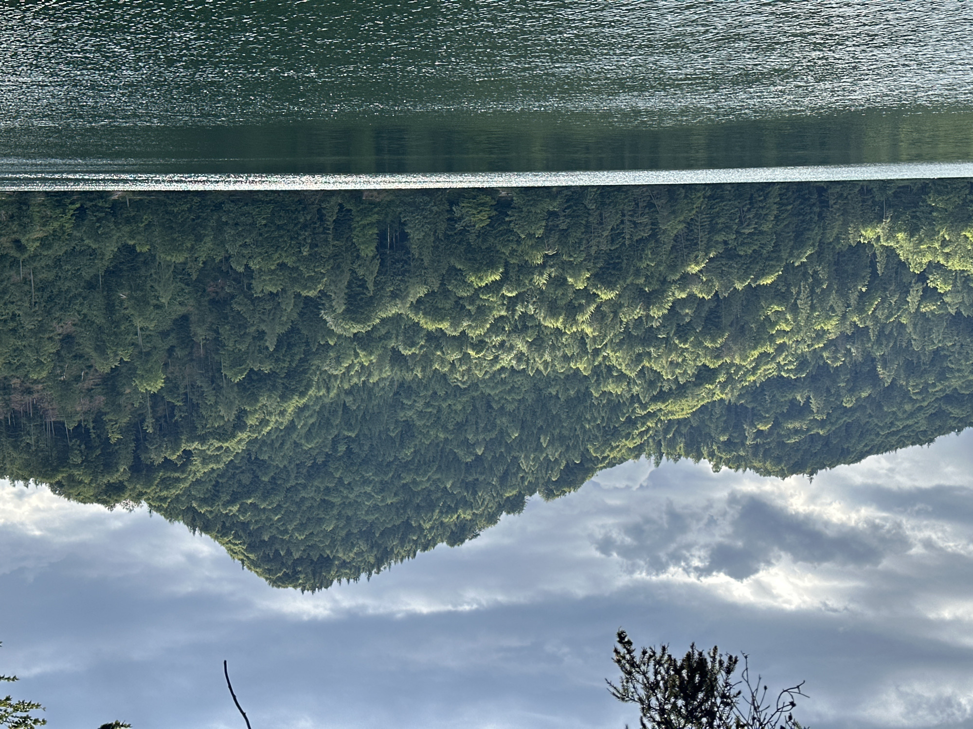 This is an outdoor, eye-level, wide shot of a serene lake with a heavily wooded mountain in the background. The water in the foreground is dark green with ripples and reflections of the sky. The mountain is covered in dense, dark green trees that extend all the way to the water's edge. The sky is cloudy with patches of sunlight breaking through, creating a dramatic contrast. The scene appears to be natural and undisturbed. This location is Kaiserklause, Kiefersfelden, Germany. There are no people or animals visible in the image.