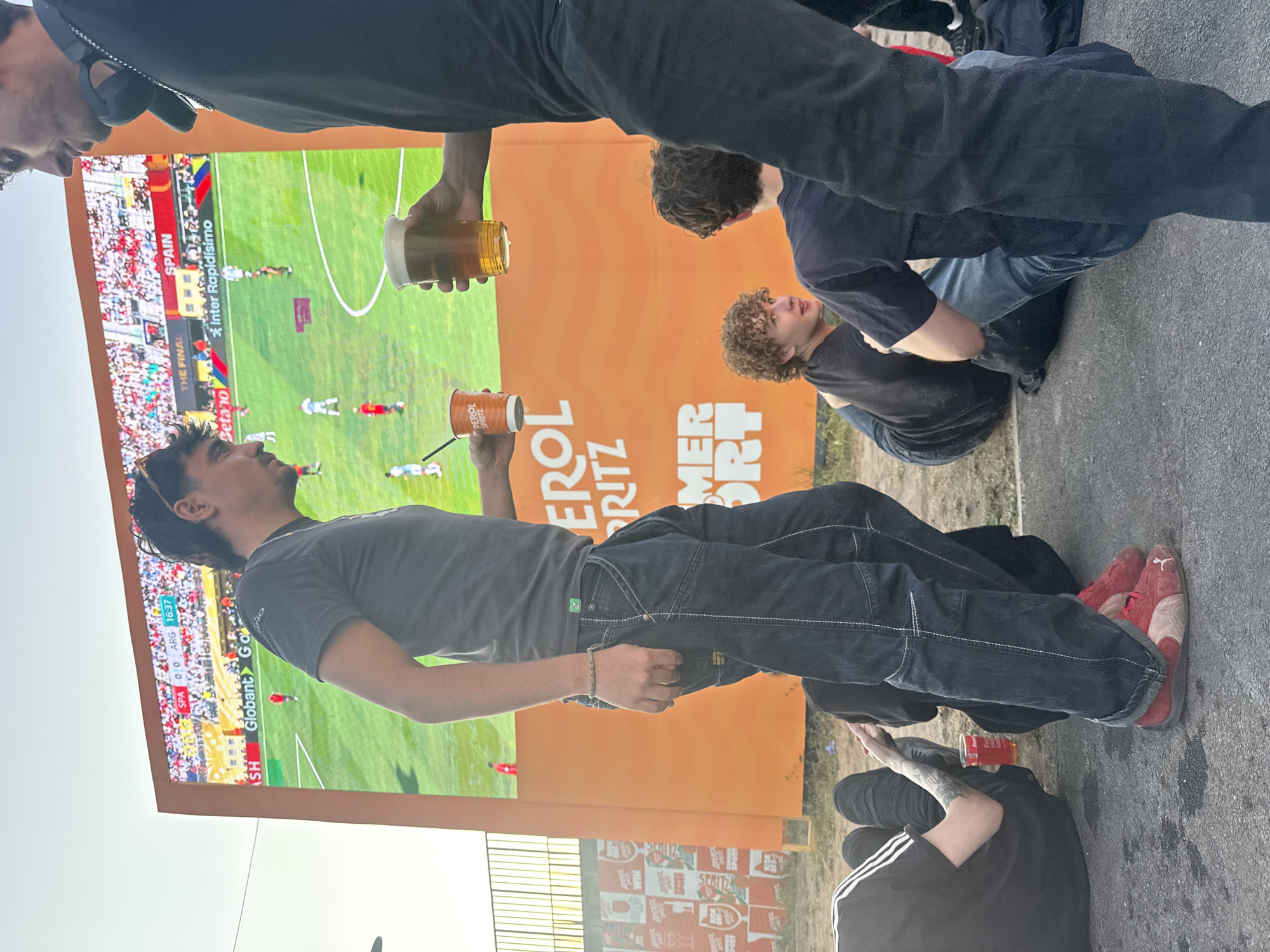 In an outdoor urban setting, a group of young adults gathers to watch a soccer match displayed on a large LED screen. The screen shows a game between Spain and Argentina, with the score currently tied at 0-0. The atmosphere appears casual and social, with some individuals holding drinks. The setting includes a prominent orange advertisement with the words "SUMMER SPORT" and "APEROL SPRITZ" visible. The ground is asphalt, and a metal fence can be seen in the background. The time of day appears to be late afternoon or early evening, given the lighting conditions.