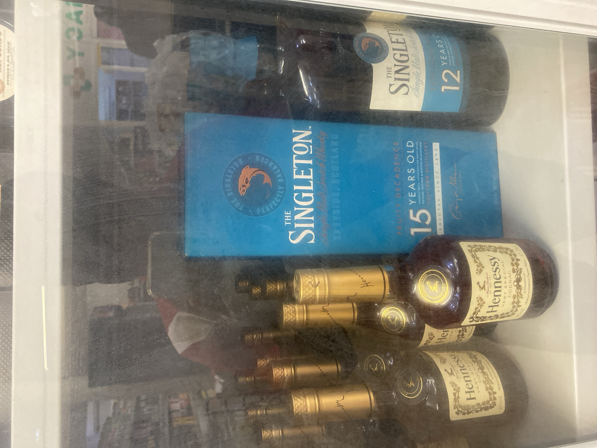 The image shows a glass-fronted refrigerated display case containing various bottles of alcoholic beverages. Prominently displayed in the center is a bright blue box for "The Singleton" single malt Scotch whisky, stating "15 YEARS OLD" and "FRUITY DECADENCE". To the right of the box is a bottle of "The Singleton" 12 Years Old Single Malt Scotch Whisky. Several bottles of Hennessy Very Special Cognac are visible on the left and in the foreground, distinguished by their amber liquid and gold foil caps. The environment appears to be a commercial establishment, likely a liquor store or supermarket, with shelves and other products visible in the blurred background, suggesting it is located in the city of Ikorodu, Nigeria. The lighting is ambient, and there are reflections on the glass, indicating it is an indoor setting. No individuals are clearly discernible in the image, though a blurred figure is visible in the reflection on the glass.