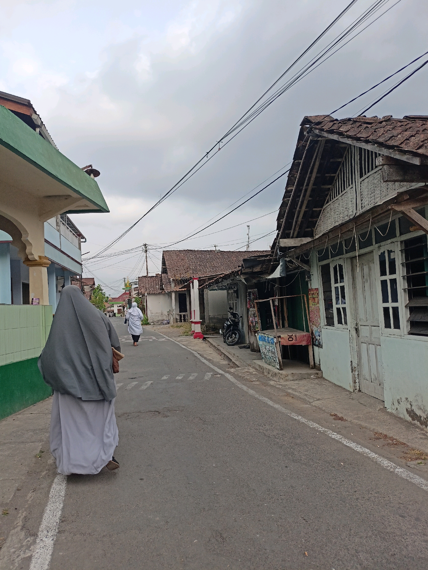 The image depicts a street scene in Bandongan, Indonesia, on an overcast day. In the foreground, a person wearing a gray hijab and a long white skirt walks away from the camera down the center of an asphalt road. The road is lined with buildings on both sides. On the left, a light blue and green building with ornate architectural details is visible. On the right, several older, weathered structures with tiled roofs and wooden elements line the street, some appearing to be shops or residences. One building on the right has a light blue facade with white-trimmed windows and doors. A motorcycle is parked on the side of the road in front of one of these buildings. In the distance, more buildings and utility poles with numerous electrical wires are visible. Another person, also wearing a white hijab, is walking further down the street on the right side. The overall impression is of a quiet, residential or commercial street in a small Indonesian town.