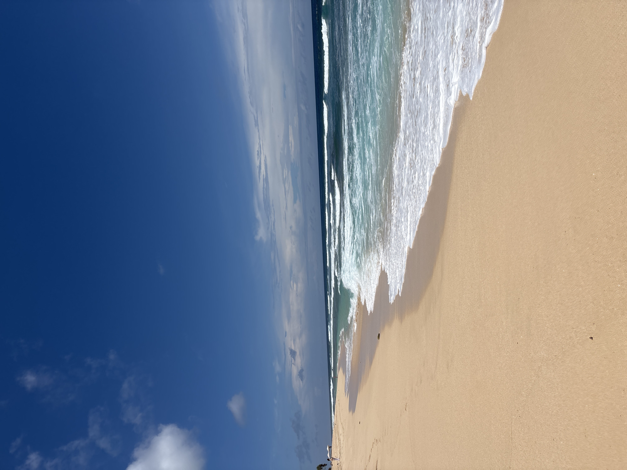 The image depicts a beach scene in Pecatu, Indonesia. The sky is blue with scattered clouds. The ocean meets the shore with crashing waves, showcasing a gradient of turquoise to white foam. The sand is a light tan color, occupying a significant portion of the frame. In the lower left, there are some trees and a few people in the far distance.
