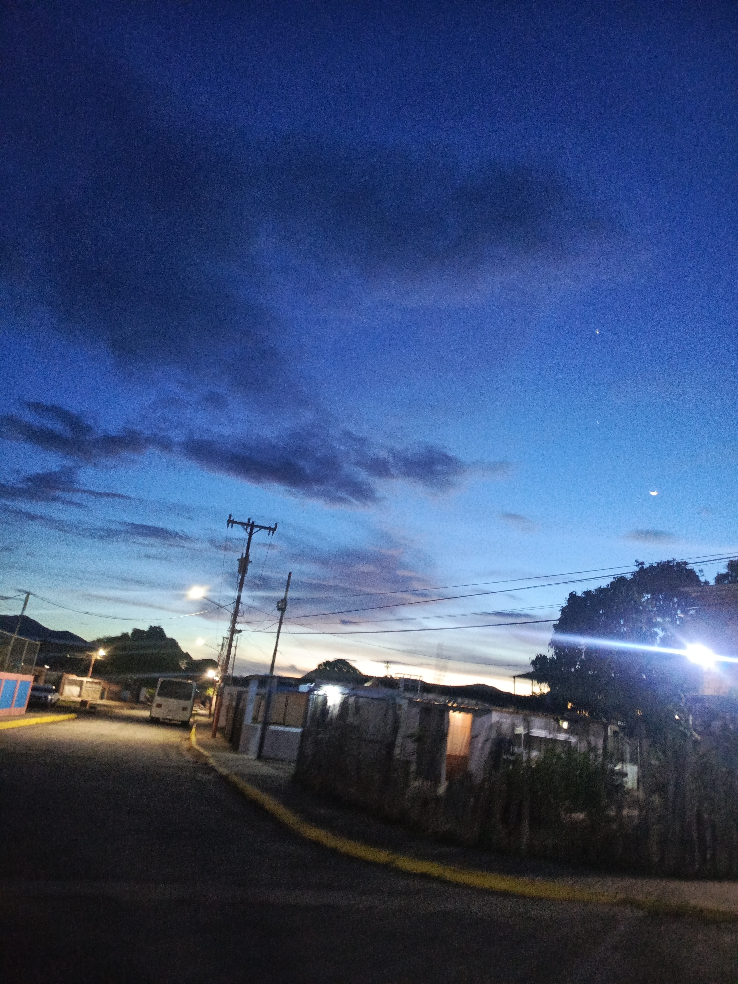 The image depicts a twilight scene on a street in Juan Griego, Venezuela. The sky is a deep blue with scattered dark clouds and a faint crescent moon visible. On the right side of the street, there are several modest dwellings with corrugated metal roofs and walls. Some lights are visible inside these structures. A large tree with illuminated foliage is also present on the right. A utility pole with electrical wires spans across the middle of the frame. On the left side of the street, a white van is parked, and further down the road, there are hills or mountains in the distance. The street itself is paved and dark, with a yellow stripe marking the edge of the road. The overall impression is of a quiet residential area at dusk.