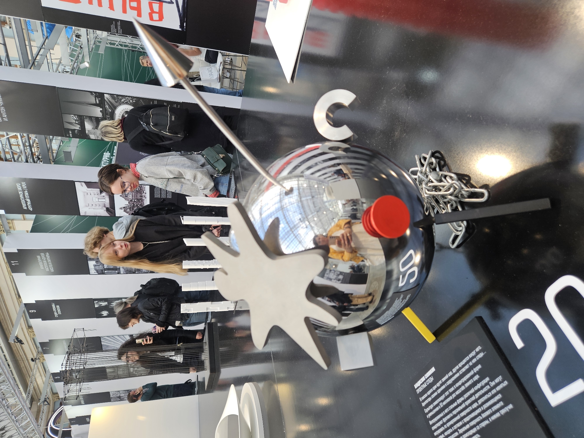 Here is a description of the image:
**Scene**: The image shows a display at an exhibition, possibly about architecture or design. There are people viewing panels behind the display.
**Objects**:
* **Display**: Features a black surface with text and various geometric elements.
* A chrome sphere with a red button and the number "50" is prominent.
* A large, star-shaped piece pierces the sphere with a spear.
* The letter "C" is another prominent shape.
* Fragments of a silver chain
* The number "20" is displayed in large white text.
* **Panels**: These display photographs of architecture and related information.
**People**:
* The reflections in the chrome sphere show people, possibly visitors to the exhibition.
**Actions**:
* People are looking at the displays.
* Some are looking down at the display and seem to be contemplating it.
**Environment**:
* The location is likely an exhibition hall in Moscow, Russia. This is inferred from the Russian text on the display and panels.
* The lighting is bright, typical of an exhibition space.