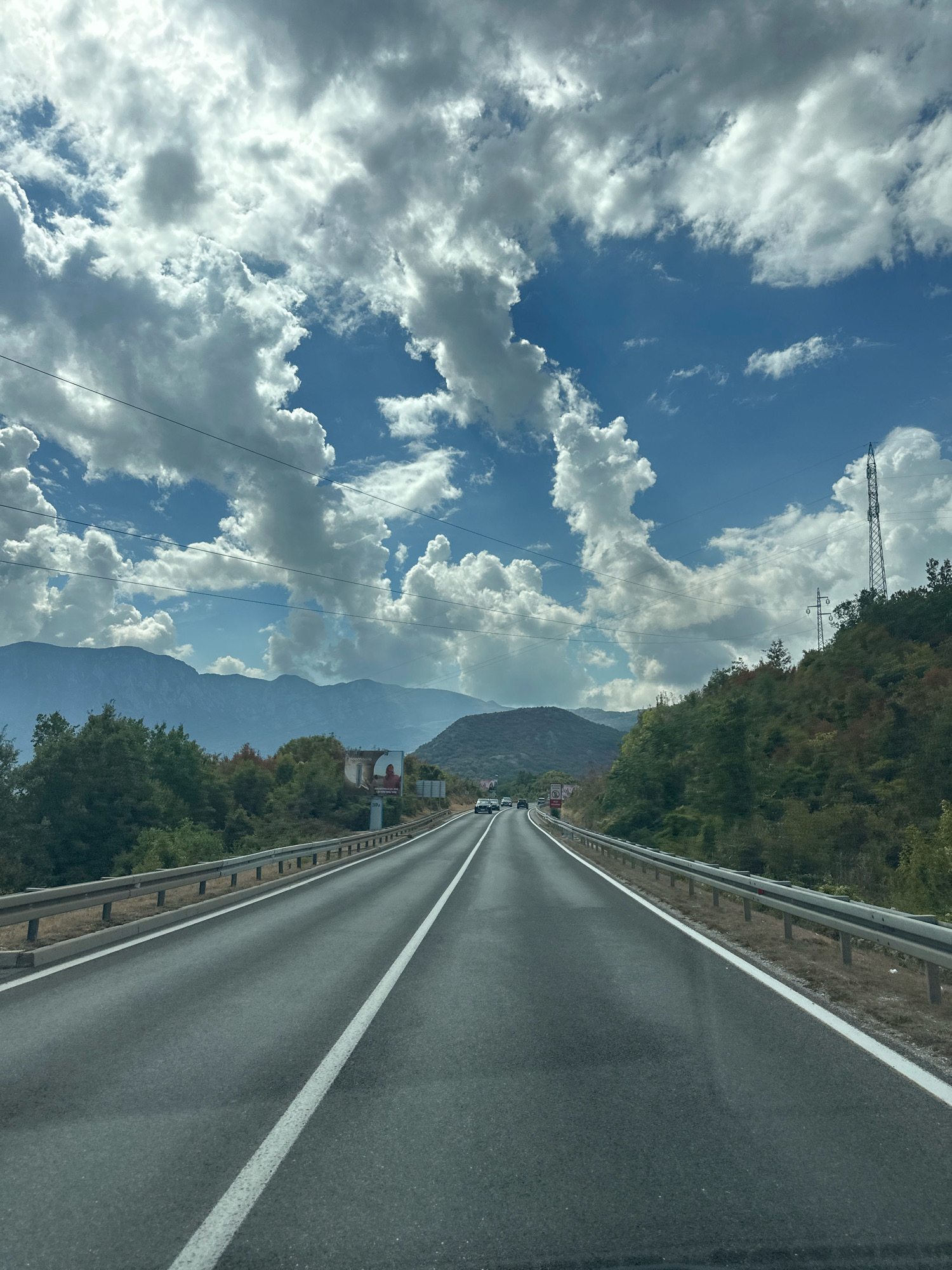 A drone shot captures a paved road stretching into the distance, flanked by guardrails and rolling green hills. Majestic mountains rise in the background under a vast sky filled with dramatic clouds. The scene appears to be the outskirts of Sotonići, Montenegro, a place where nature's grandeur meets the open road.