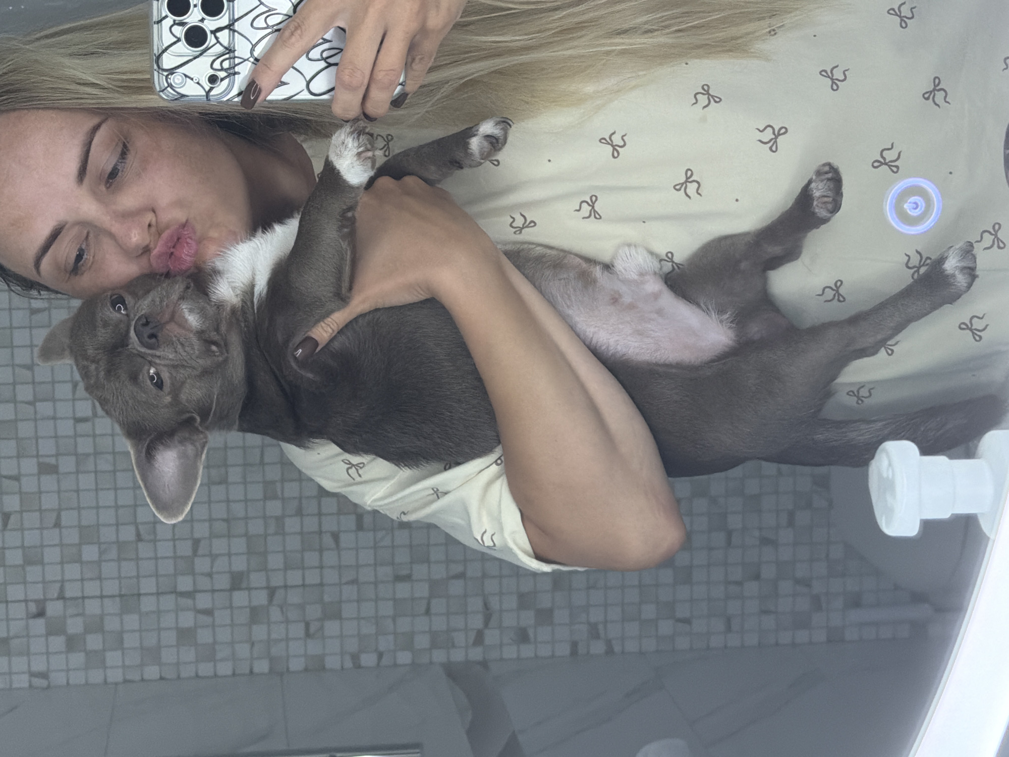 In a bathroom, a woman with blonde hair holds a gray and white dog, kissing it. The woman is taking a selfie with a phone in her right hand. The dog is positioned vertically, held by the woman's arms. The background features a tiled wall. The scene is illuminated by artificial light. The exact location of Votkinsk, Russia is not discernible from the image.