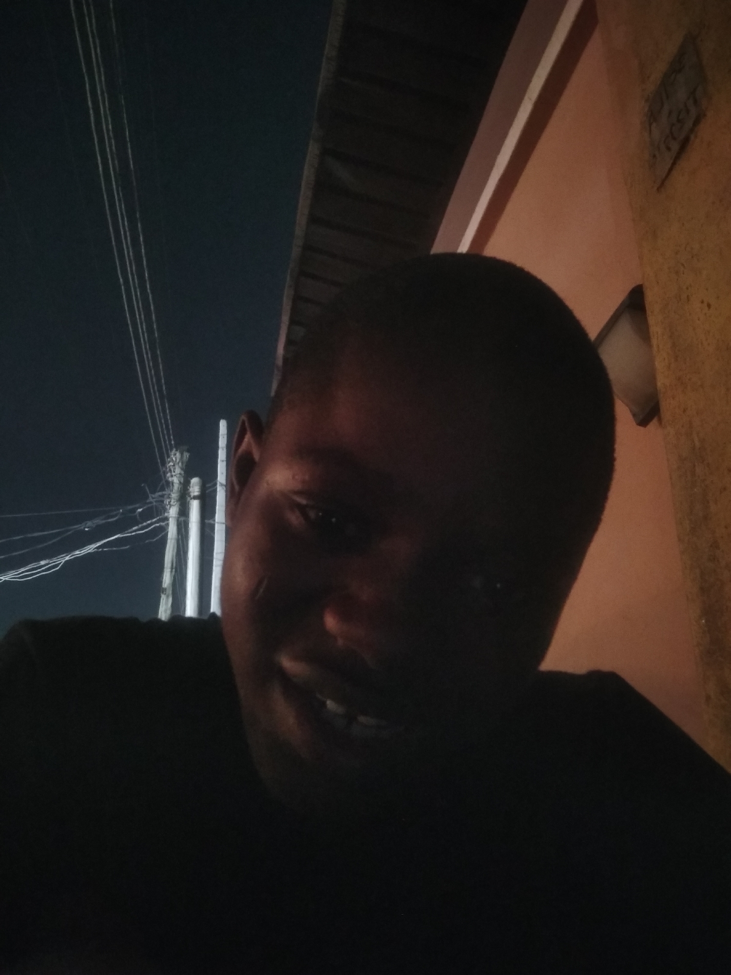 This image captures a close-up selfie of a person, likely male, against a nocturnal outdoor backdrop in Shangisha, Nigeria. The individual, with dark skin and closely cropped hair, occupies the lower right portion of the frame. Their face is largely silhouetted due to low light conditions, but a clear smile is visible, revealing teeth, suggesting a cheerful or content mood. They are wearing a dark-colored shirt.
In the background, visible through the darkness, are distinct features of an urban or suburban setting. To the left, numerous power lines or utility cables stretch across a dark sky, converging towards a utility pole that is partially obscured by the person's head. The white lines of the cables stand out against the deep blue-black of the night sky. To the right of the person, a building wall is visible, painted in a faded peach or light orange color with a darker trim above. A small, square-shaped wall-mounted light fixture is visible on this wall, though it does not appear to be illuminating the immediate area.
Faint text is discernible on a sign affixed to the wall in the upper right corner. Though largely illegible due to resolution and lighting, parts of words like "NOT BE" and "POSIT" can be made out, suggesting a phrase such as "DO NOT DEPOSIT" or similar. The overall impression is an everyday scene in a residential or commercial area during nighttime, characterized by common infrastructure elements and a friendly human presence.