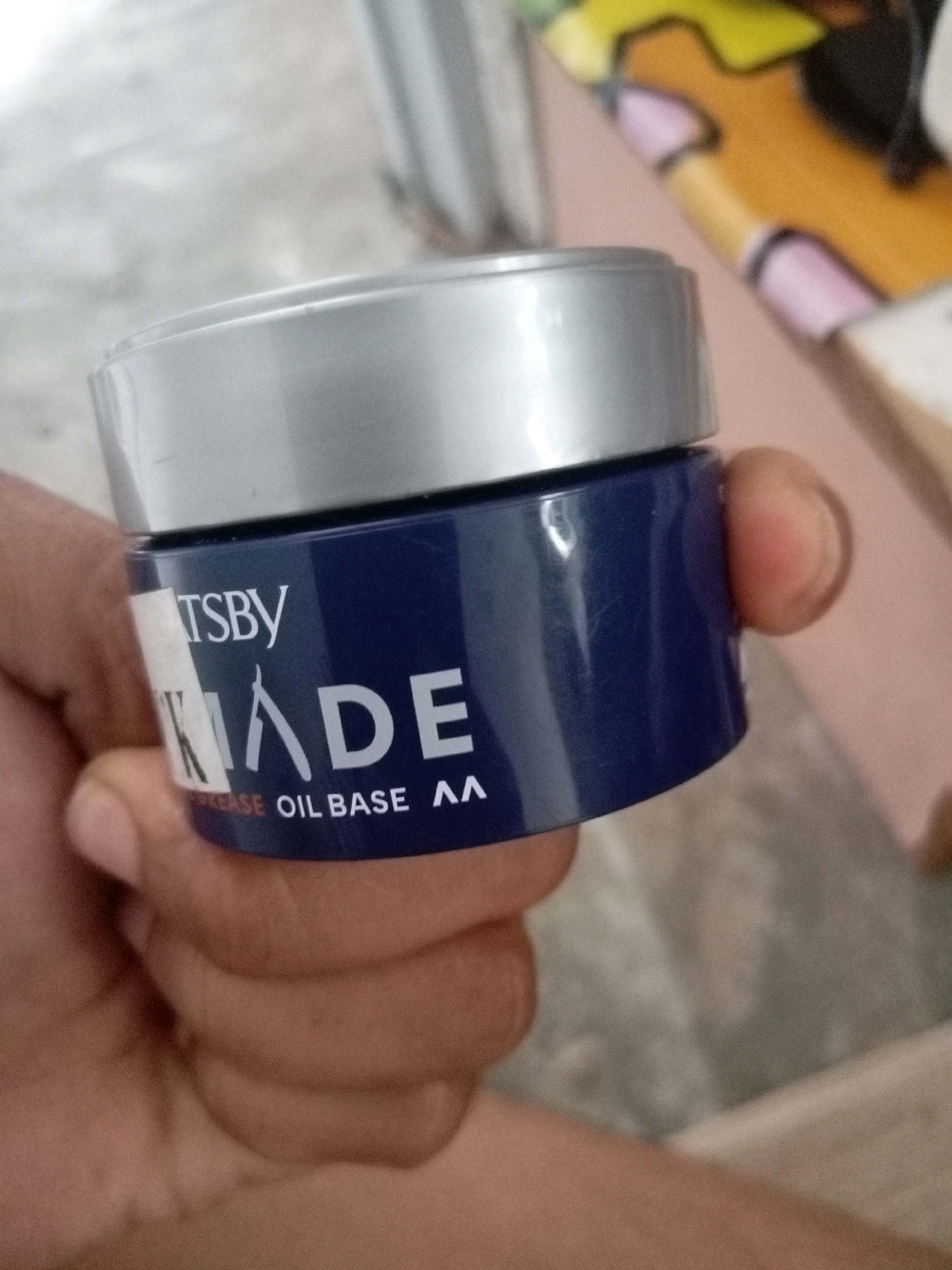 A hand is holding a dark blue jar with a silver lid, likely hair pomade. The jar has white lettering on it. The word "TSBY" is visible at the top, followed by a stylized "K" and the word "ADE" below. Underneath, the words "GREASE OIL BASE AA" are also visible. The background is blurred and appears to be indoors.