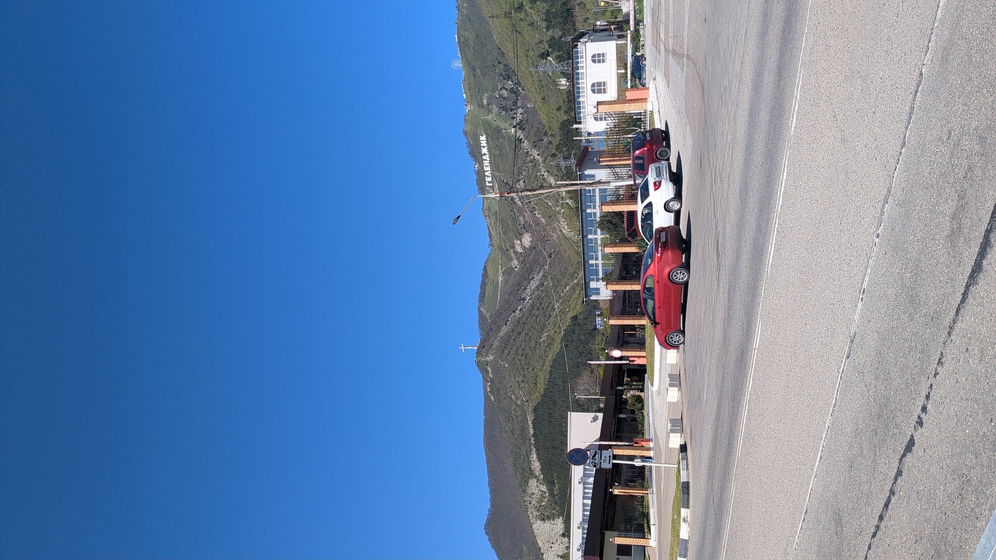 The image, correctly oriented, depicts a clear, sunny day in Gelendzhik, Russia. In the background, a large, green-brown mountain rises steeply, partially covered in vegetation. Prominently displayed on the mountainside are large white letters spelling "ГЕЛЕНДЖИК" (Gelendzhik). Higher up on the mountain, a large white cross is visible, and near the summit, what appears to be a Ferris wheel can be discerned. Utility poles and overhead lines traverse the scene in front of the mountain.
In the mid-ground and foreground, an asphalt road runs horizontally, marked with distinct white lines. Three cars are parked along the right side of the road: a red sedan in the front, a white sedan, and a dark-colored SUV. Adjacent to the road, several multi-story buildings with balconies and some brick facades line the street. A concrete curb or sidewalk with light-colored bollards separates the buildings from the road. The sky above is a brilliant, cloudless blue.
The scene captures a static street view, likely in a coastal or mountainous resort town. The time of day is bright daylight, suggesting mid-morning to late afternoon. The visible text "ГЕЛЕНДЖИК" clearly identifies the location, and the mountain with its landmarks serves as a prominent geographical cue. No people or specific activities are clearly visible.