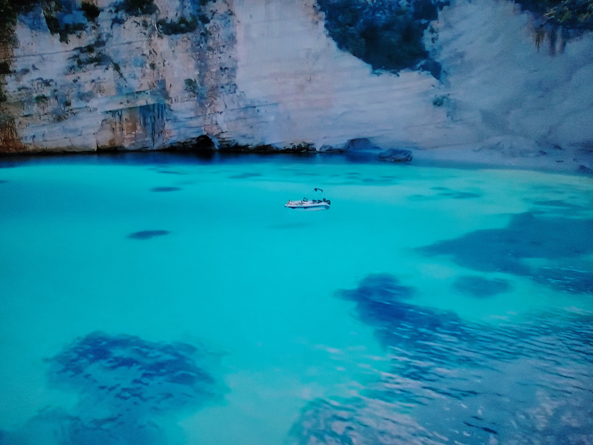 A small white boat with a dark canopy and outboard motor is centered in clear turquoise water. The boat is positioned in front of a rugged, light-colored cliff face that recedes into the distance. The water is shallow enough in places to reveal dark, submerged rocks or reef formations. The overall scene depicts a tranquil cove with a boat at anchor or slowly moving. The location is not Huntingdonshire, United Kingdom, but rather appears to be a coastal environment, possibly a Mediterranean bay or a tropical island setting, given the water clarity and rock formations. There are no people visible on the boat or in the water.