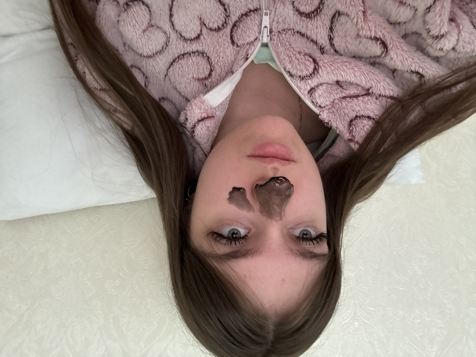 The image shows a young person lying down, captured from an overhead perspective, creating an upside-down appearance. The individual has long, dark brown hair that frames their face and spreads out on a light-colored surface. They are wearing a pink, fuzzy jacket or robe with a distinct heart or swirl pattern and a white zipper, which is partially open, revealing a light green or mint-colored garment underneath.
A dark, possibly charcoal or mud-based, facial mask is applied to the person's nose and forehead area. Their eyes are wide open and directed towards the viewer, with prominent, long eyelashes. Their lips are slightly parted. To the left of the person's head, a white pillow or sheet is visible. The background on the right and at the bottom features a light-colored surface with a subtle embossed or patterned design, suggesting a duvet cover or textured wallpaper in an indoor setting, likely a bedroom.
The activity taking place appears to be a self-care or relaxation routine involving a facial mask. No text is visible in the image.