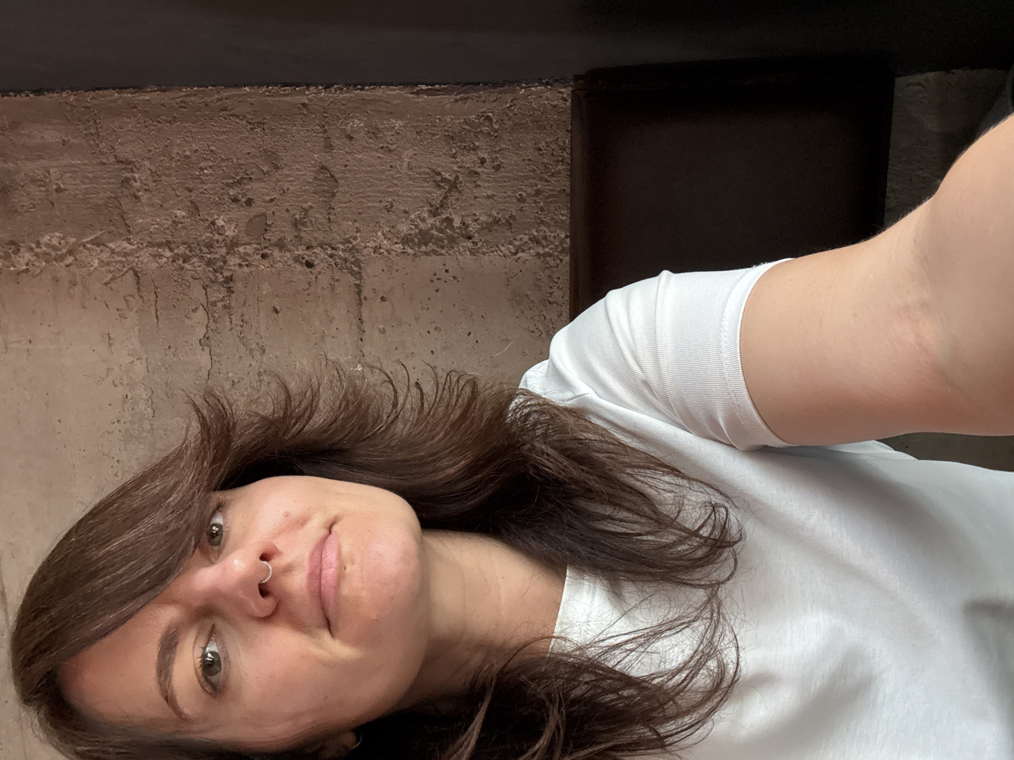 This vertical selfie captures a woman indoors in Podgorica, Montenegro, looking directly at the camera with a calm, slightly smiling expression. Her head is tilted to her left, and her body is angled towards the upper right of the frame.
The subject is a woman with medium-length, wavy, dark brown hair that falls around her face and over her shoulders. She has dark eyes and fair skin, with a small, silver-toned hoop piercing visible in her right nostril. She is wearing a simple white short-sleeved t-shirt.
The background features a textured, brownish-grey wall with a rough, uneven surface, resembling exposed concrete or stucco. Above this wall, a dark, horizontal band, possibly a ceiling or an architectural element, spans the top of the image. To the right, another dark, vertical shape suggests a corner or a piece of furniture. The lighting is soft and diffuse, indicating an indoor setting without strong natural light. No text is visible.