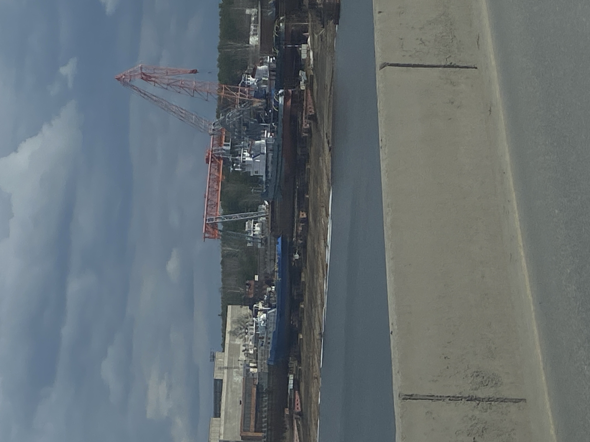 The image shows a crane in what appears to be an industrial or construction site. In the background are several buildings, possibly part of a factory or storage facility. The sky is overcast with heavy clouds. The foreground contains parts of a concrete structure. The location is Chaikovsky, Russia. There are no visible people.