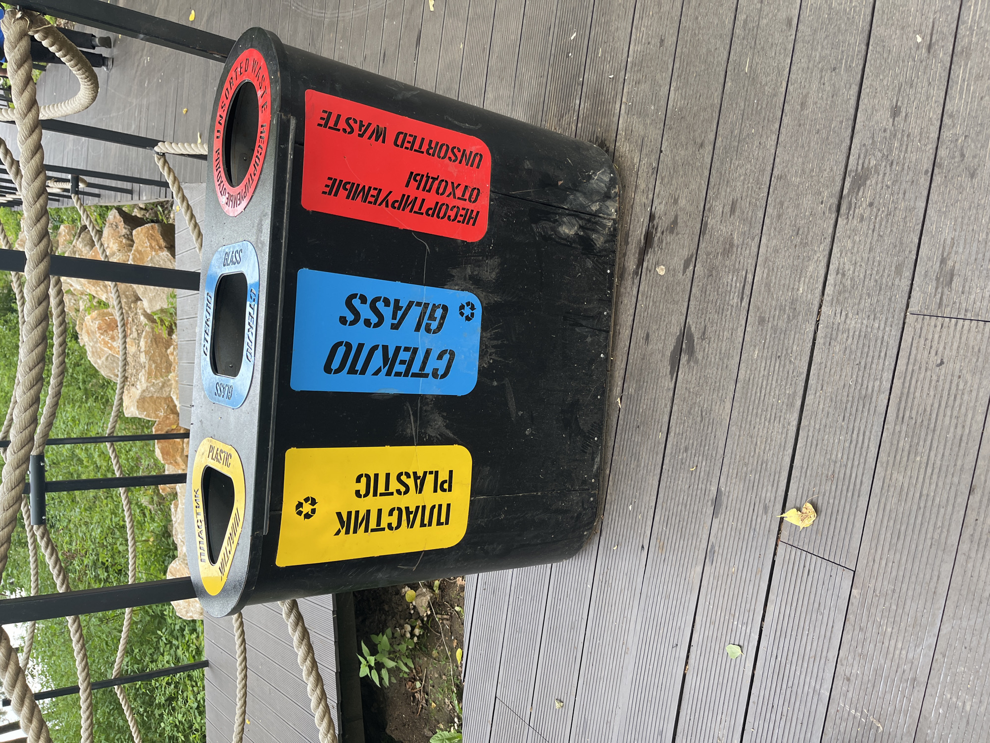 A three-compartment recycling bin is situated on a wooden deck outdoors. The bin is black and features yellow, blue, and red labels, each indicating a different type of waste. The yellow compartment is labeled "ПЛАСТИК PLASTIC" with a recycling symbol. The blue compartment is labeled "СТЕКЛО GLASS". The red compartment is labeled "НЕСОРТИРУЕМЫЕ ОТХОДЫ UNSORTED WASTE". The bin is positioned near a railing made of rope and metal poles, beyond which is greenery and some large rocks. The wooden deck has a weathered appearance, with some fallen leaves visible. The context provided about Blagoveshchensk, Russia is not depicted in the image itself.