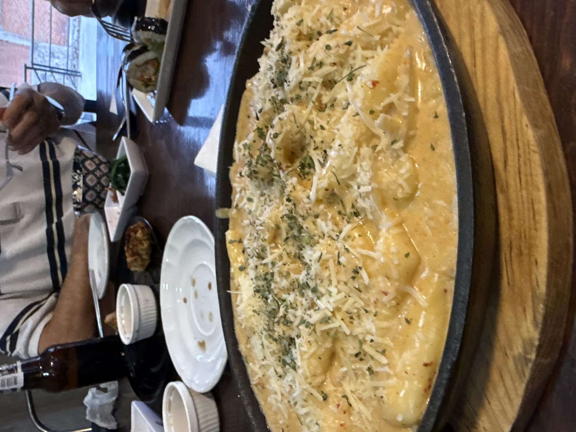 The image shows a table setting in what appears to be a restaurant. A person wearing a striped shirt is visible on the left side of the frame, using cutlery to eat. A plate of sushi is present on the table, along with small bowls of food. The main subject is a large oval dish of pasta with a creamy sauce, topped with shredded cheese and herbs. A brown beer bottle is also on the table, next to small white dishes. Based on visual context and the presence of dishes commonly found in Mexican cuisine, the location is potentially in Puebla, Mexico, although further evidence is needed to confirm the exact location.
