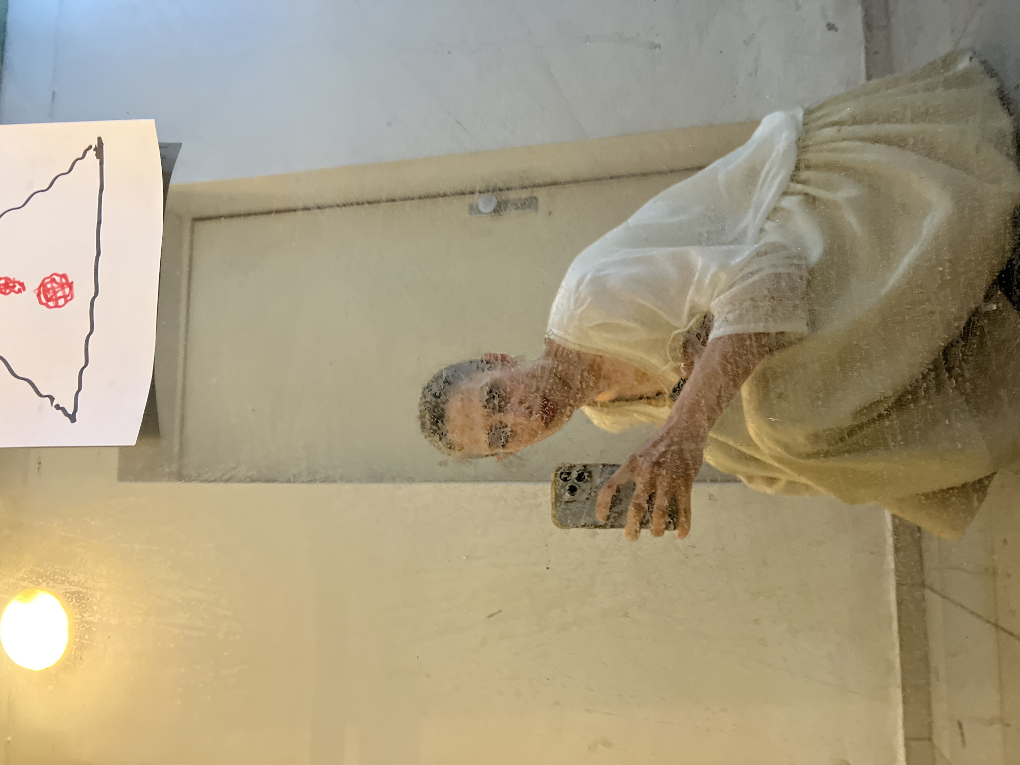 An indoor mirror selfie shows a person, likely female, with short hair, reflected in a large, somewhat smudged or wet mirror. The person is wearing a light-colored, possibly cream or white, flowing dress featuring gathered fabric details on the sleeves and skirt, suggesting a historical or bohemian style. They are holding a smartphone in their right hand, positioned to capture the reflection.
The individual's face, partially obscured by the mirror's condition, is turned towards the camera with a neutral or slightly smiling expression. To the left of the reflection, a piece of white paper is attached to the mirror, displaying a drawing with a black outline of an irregular shape and two red, scribbled circles inside. A bright, circular light source is visible in the lower left corner of the frame. The background visible in the reflection suggests plain, light-colored walls and a door frame. The mirror surface has numerous droplets or smudges, creating a hazy effect on the reflection. The scene is located in Sofia, Bulgaria. No legible text is visible in the image.