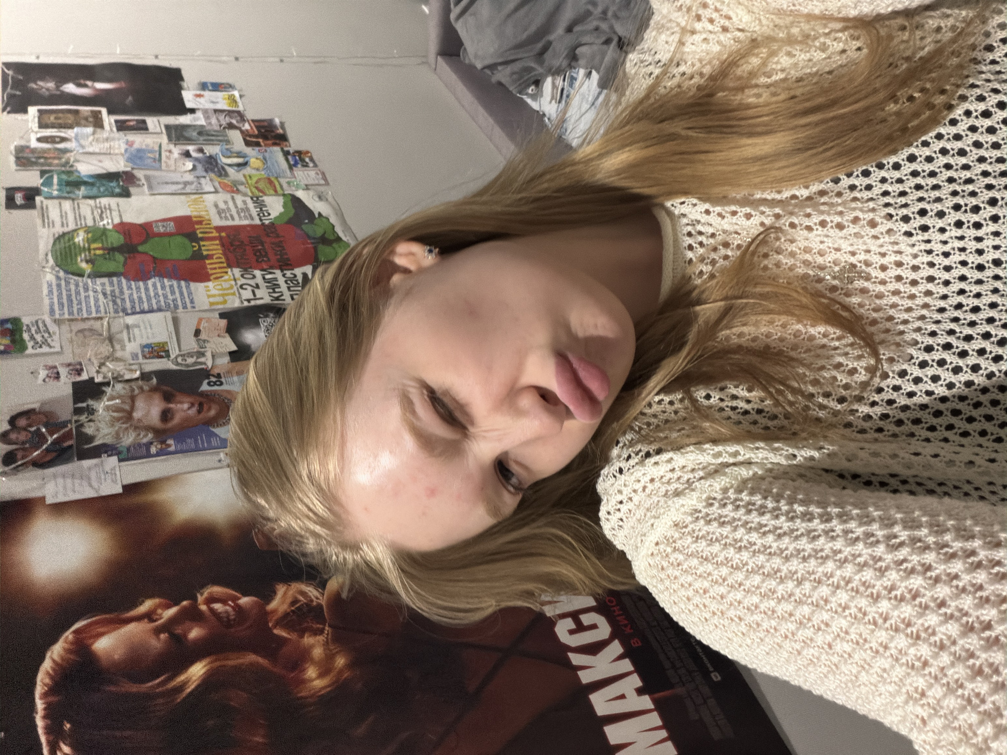 A young, light-skinned person with long, blonde hair is posing for a photo, sticking out their tongue and squinting their left eye. They are wearing a white, crocheted sweater. Behind them, a wall is covered with various posters and images. One poster prominently features the word "MAKC" in Cyrillic letters and mentions "Kino" (cinema). Another poster contains Russian text and a cartoon-like figure. The environment seems to be a bedroom or living space in a residential area. Based on the Cyrillic text on the posters, the location might be Засечное, Russia.
