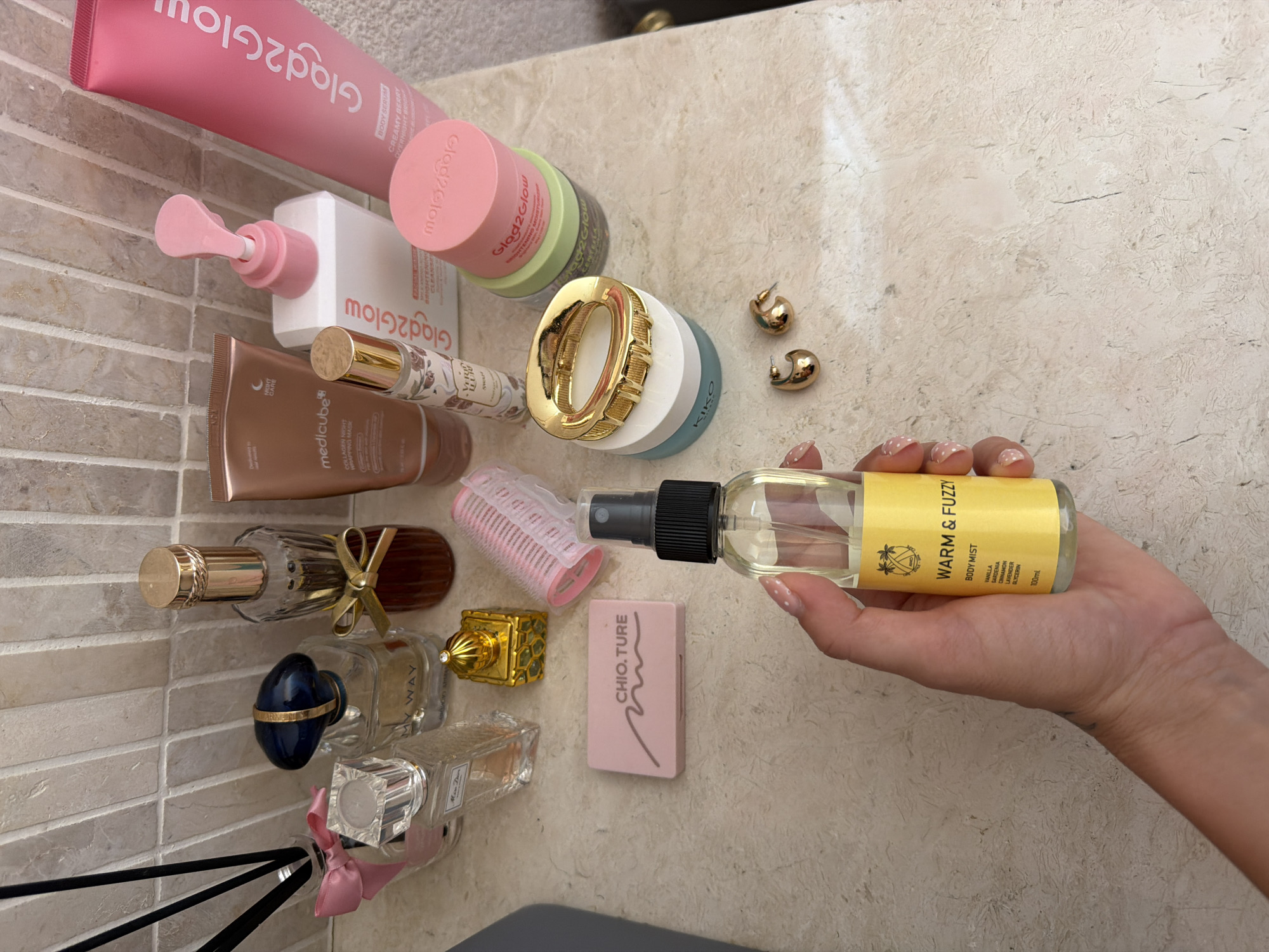 The image shows a collection of beauty products and accessories arranged on a countertop with a tiled backsplash. In the foreground, a hand is holding a clear spray bottle with a yellow label that reads "WARM & FUZZY BODY MIST." The background features several perfume bottles, skincare products, and a small pink compact. The countertop has a textured surface with subtle variations in color. The environment appears to be a domestic setting, likely a bathroom or vanity area. There is no indication of the city of Mengwi, Indonesia in the image or its contents.
