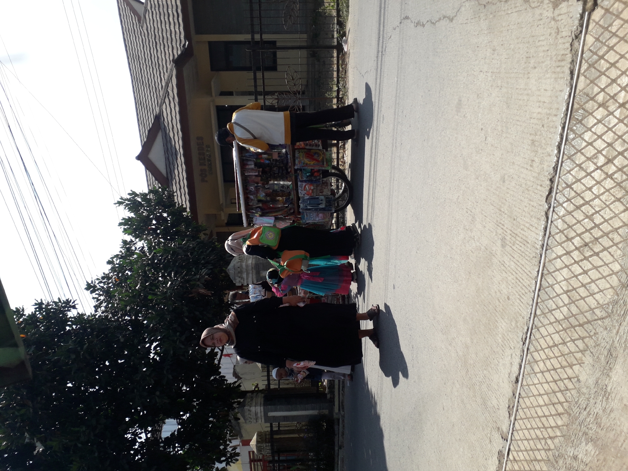 In Cankuang, Indonesia, on a paved road, a street vendor with a cart displays various items. The cart is laden with colorful goods, possibly toys or trinkets. Several individuals are near the cart. A woman in a black abaya and a headscarf stands prominently in the foreground, her shadow cast on the asphalt. To her right, a child in a colorful dress and a woman wearing a hijab are visible. Another person, a man in a yellow and white jacket and black pants, stands behind the cart, appearing to interact with it. The background shows a building with "POS KESDES" written on it, suggesting it is a village post office or community center. Trees line the street, and a metal fence runs along the property. The scene is illuminated by bright sunlight, casting distinct shadows.