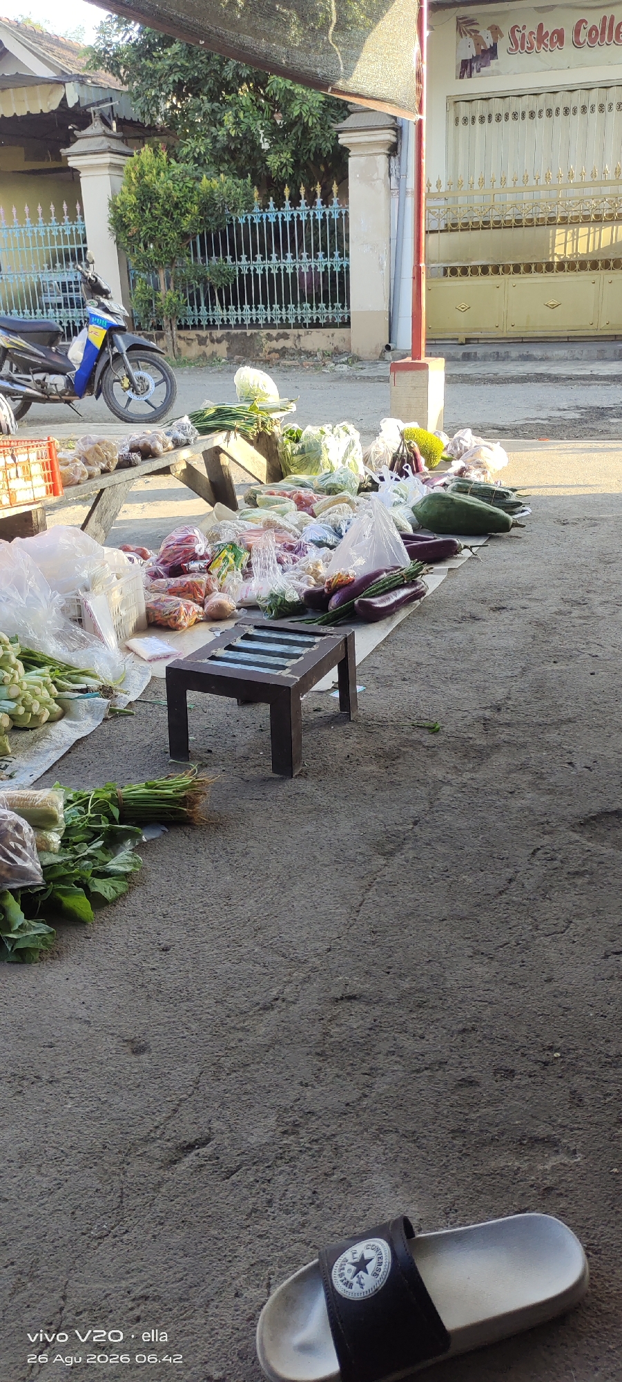 A roadside market stall in Simo, Indonesia, displays a variety of fresh produce and packaged goods under a dappled canopy of trees and netting. The stall is set up along a concrete road, with a fence and buildings visible in the background. A blue and yellow motorcycle is parked to the left, partially obstructing the view of the market.
The produce includes leafy greens like spinach and long beans, as well as eggplants, papayas, and what appear to be cabbages. Some of the items are bundled, while others are loose or packaged in clear plastic bags. A low, dark wooden stool sits in the foreground, likely for customers or the vendor. The ground is paved with concrete and shows some cracks and discoloration. A single slide sandal with a Converse logo is positioned in the lower right corner of the image. The overall scene suggests a typical morning market in a rural or semi-urban Indonesian setting.