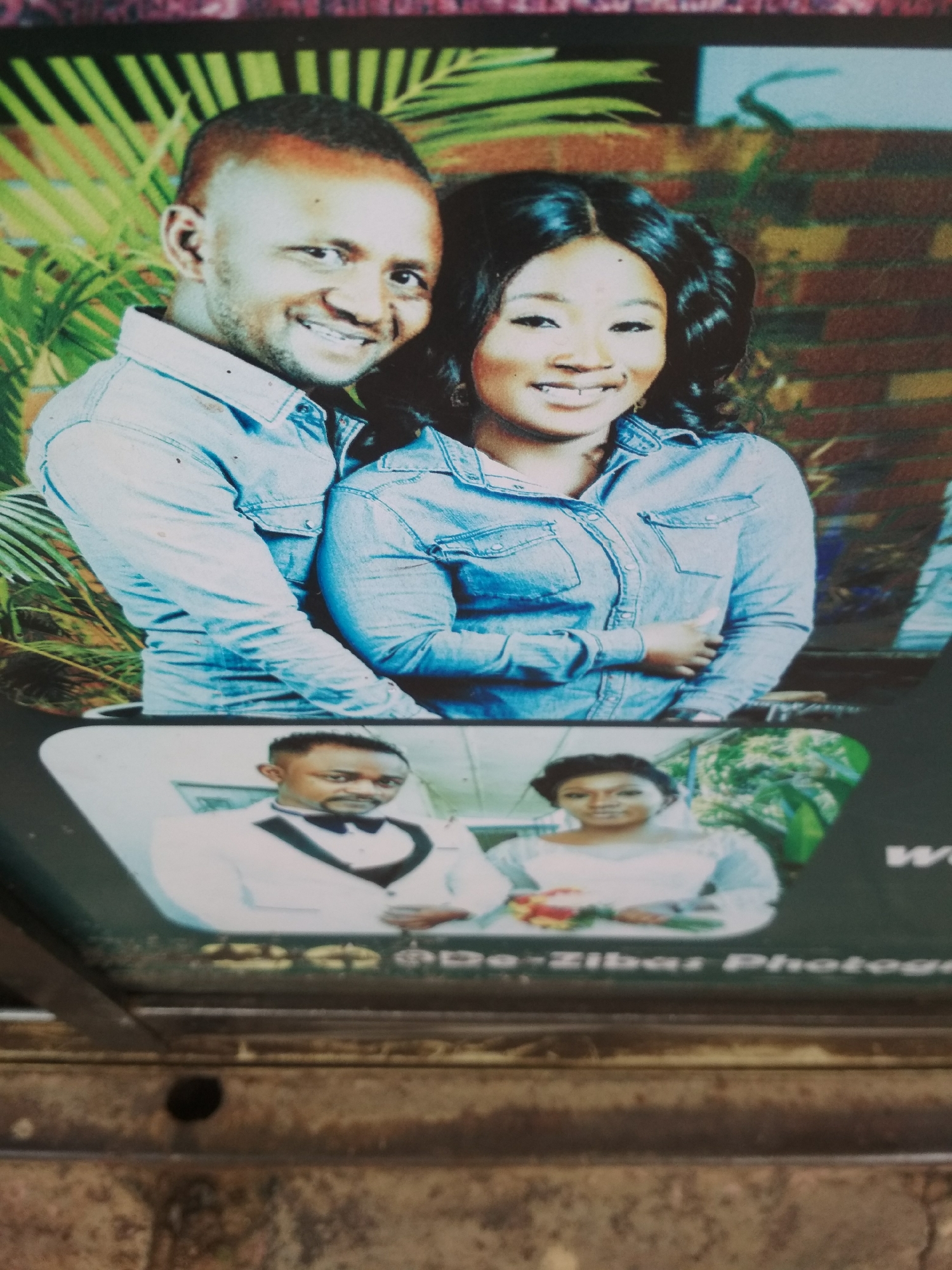 The image depicts a photograph of a couple, likely a promotional image for a photography studio. The upper portion features a man and a woman in denim shirts, smiling and embracing in an outdoor setting with green foliage and brickwork in the background. Below, there is a smaller inset with another photograph of the same couple, presumably from their wedding day. The man is in a white tuxedo with a bow tie, and the woman is in a white dress holding a bouquet. The text "De-Zibar Photography" is visible, suggesting this is an advertisement. The overall presentation is on a dark surface, possibly a counter or display stand. There is no discernible information available in the photo to determine its specific location in Jos, Nigeria.