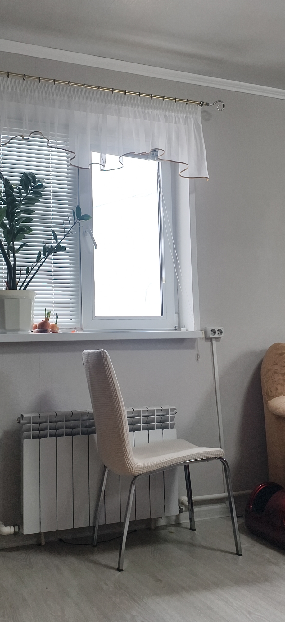 The image shows an interior setting, possibly a room in Фоки, Russia. A chair with a light beige fabric seat and chrome legs stands in front of a radiator, which is white. Behind the chair is a window with closed blinds and a white sheer curtain trimmed with a thin brown line. On the windowsill sits a potted plant. An electrical outlet with a long white extension cord is to the right of the window. To the right of the chair and extension cord, a tan-colored piece of furniture, possibly a chair or sofa, is partially visible. A red object is also visible near the right edge of the image, partially obscured by the chair's legs. The walls are painted a light grey. The floor is a light grey laminate or vinyl. The scene appears to be indoors and stationary, suggesting a domestic or residential location.
