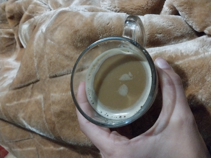 A hand holds a glass mug filled with a coffee-colored beverage. The drink has a thin layer of foam along the edge and two distinct light-colored spots in the center. The background is a tan blanket with a textured, quilted pattern. The blanket's soft, plush fabric suggests a cozy and warm setting. The angle is slightly above, looking down into the mug.