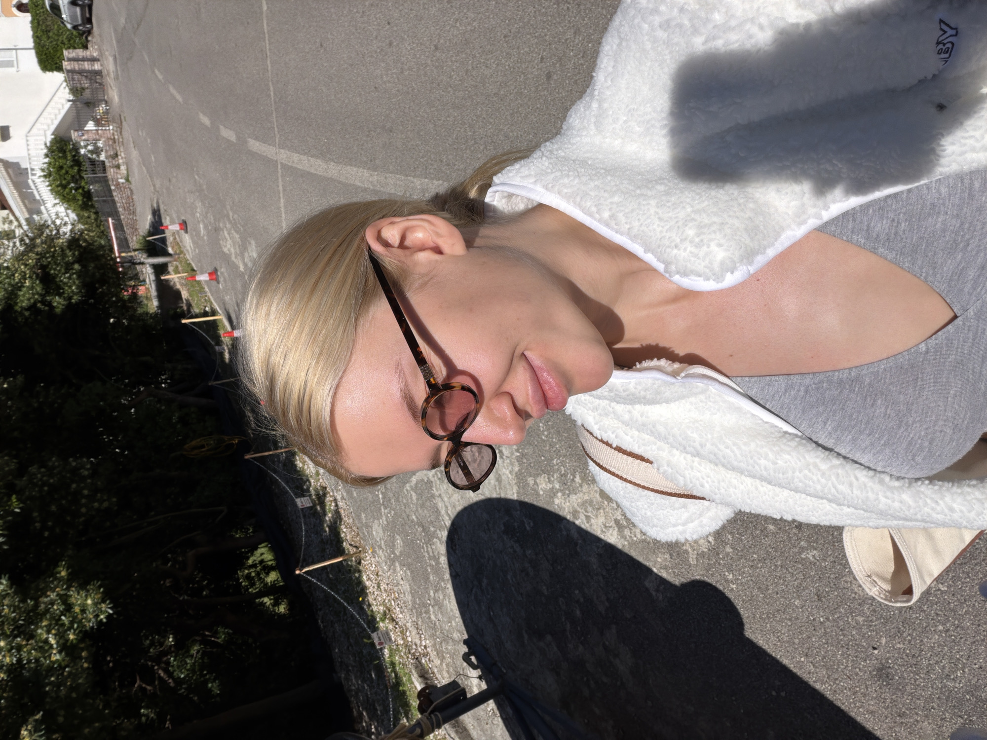 The image shows a light-skinned person with blonde hair wearing sunglasses, a grey top, and a white fleece jacket. The person is likely standing or sitting on a paved surface, possibly a road or parking lot. To the left of the person is greenery and buildings, suggesting an urban or suburban environment. The scene is outdoors in daylight. The location is possibly Nerin, Montenegro.