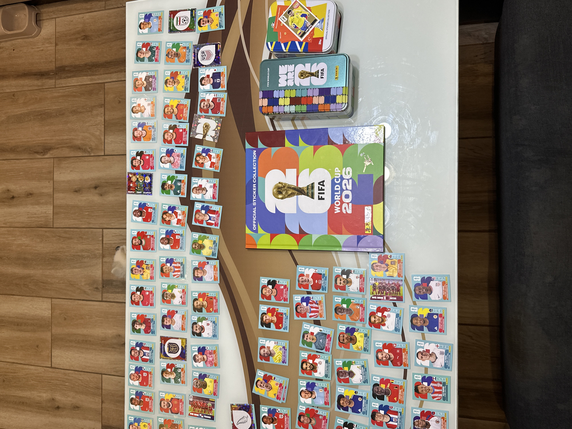 The image displays a collection of FIFA World Cup 2026 stickers and related merchandise. A sticker album and two tin boxes, one blue and one green, are prominently featured on a white surface. The tin boxes contain various stickers, including one with the text "WE ARE ONE FIFA" and another showcasing a soccer player. Scattered around these items are numerous individual stickers, each featuring a portrait of a soccer player with their name and country. These stickers are arranged in rows, suggesting a hobby or collection. The background consists of a wooden floor. The scene is indoors, and there are no people or animals visible in the immediate vicinity. There is no indication of the city of Prisovo, Bulgaria in this image.