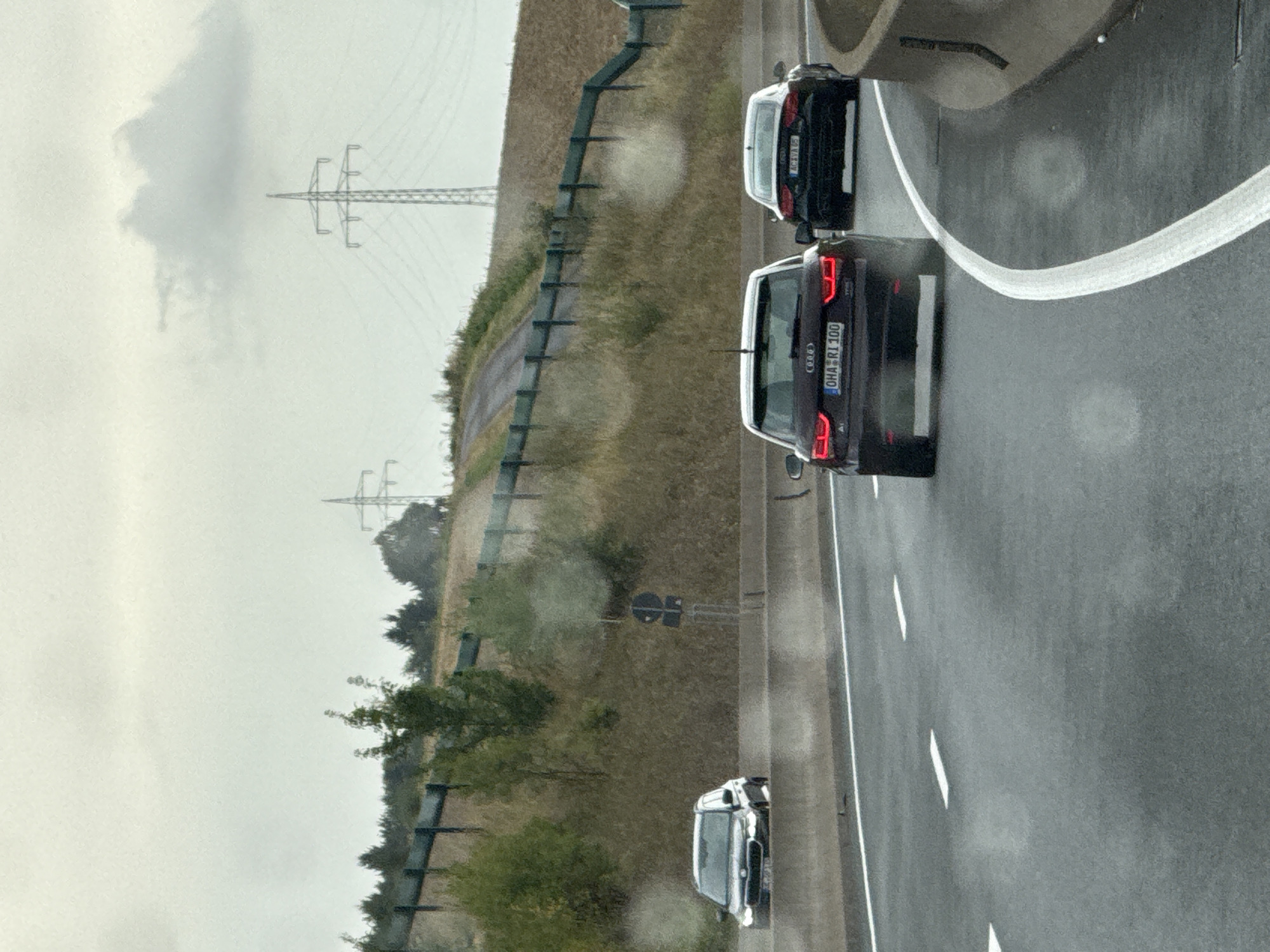The image depicts a highway scene under a cloudy sky in Bad Lauterberg im Harz, Germany. Three cars are visible on the road. On the left, a dark-colored hatchback is seen from the front-left, slightly angled. In the center, a dark red Audi hatchback is observed from the rear, with its license plate reading "OHA RI 100". To the right of the Audi, a black Audi sedan is visible from the rear, with its license plate displaying "AC XVA 95". The cars are on a multi-lane highway with concrete barriers and white lane markings. The surrounding environment includes a grassy embankment with trees and foliage, and in the background, power line towers are prominent against the sky. The road surface appears wet, suggesting recent rain.