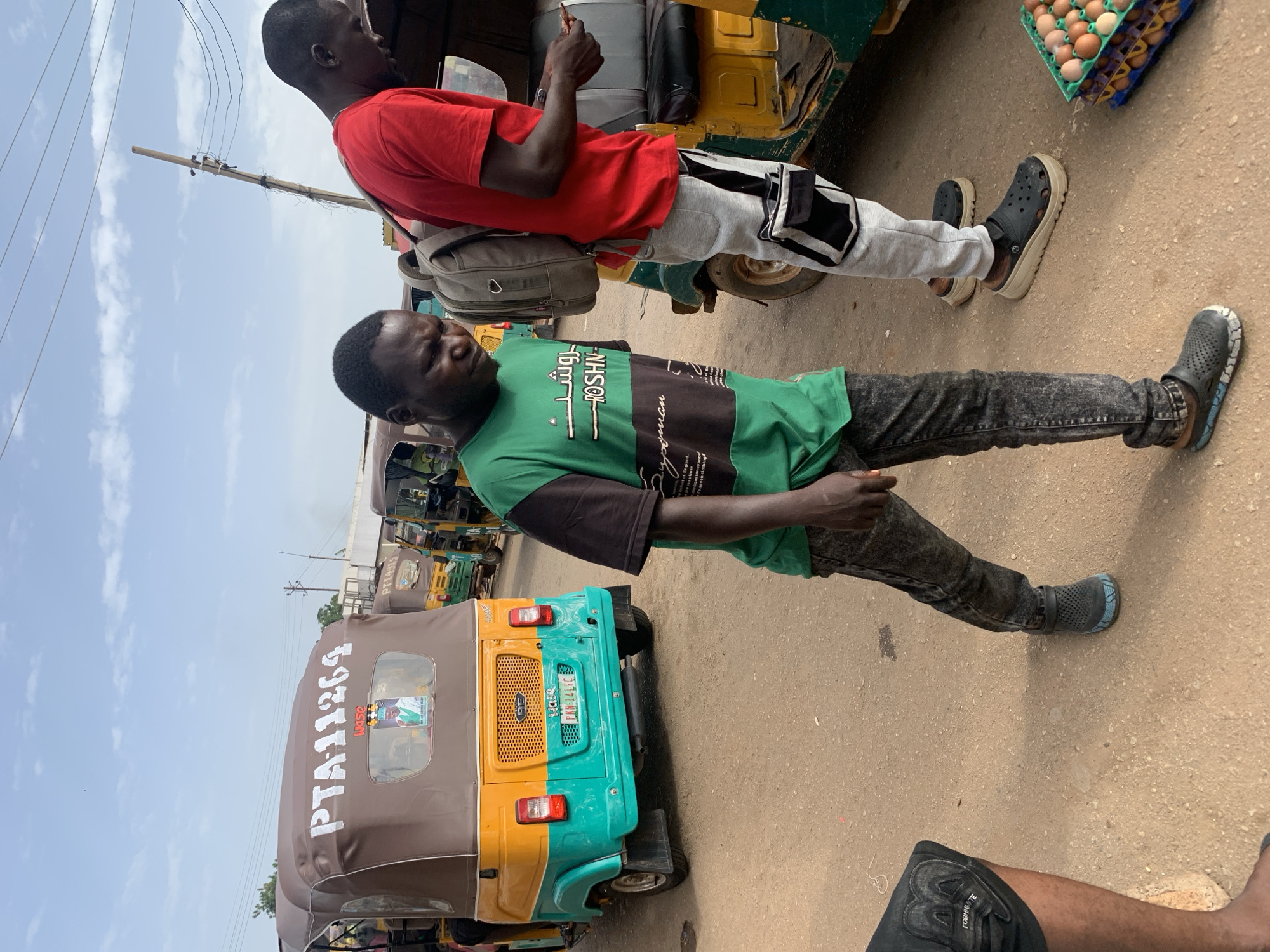 In Gura Top, Nigeria, two men are standing in a dusty street beside several auto-rickshaws. The man on the left wears a green and black t-shirt with Arabic writing on it, dark denim jeans, and black clogs. The man on the right wears a red t-shirt, light gray sweatpants with black accents, and black clogs. He has a backpack on and is looking at his phone. In the background, an auto-rickshaw is visible with the license plate PTA-11264 and a picture of a man inside. Another auto-rickshaw to the right has a crate of eggs on the ground beside it. The sky is partly cloudy and the ground is dirt.