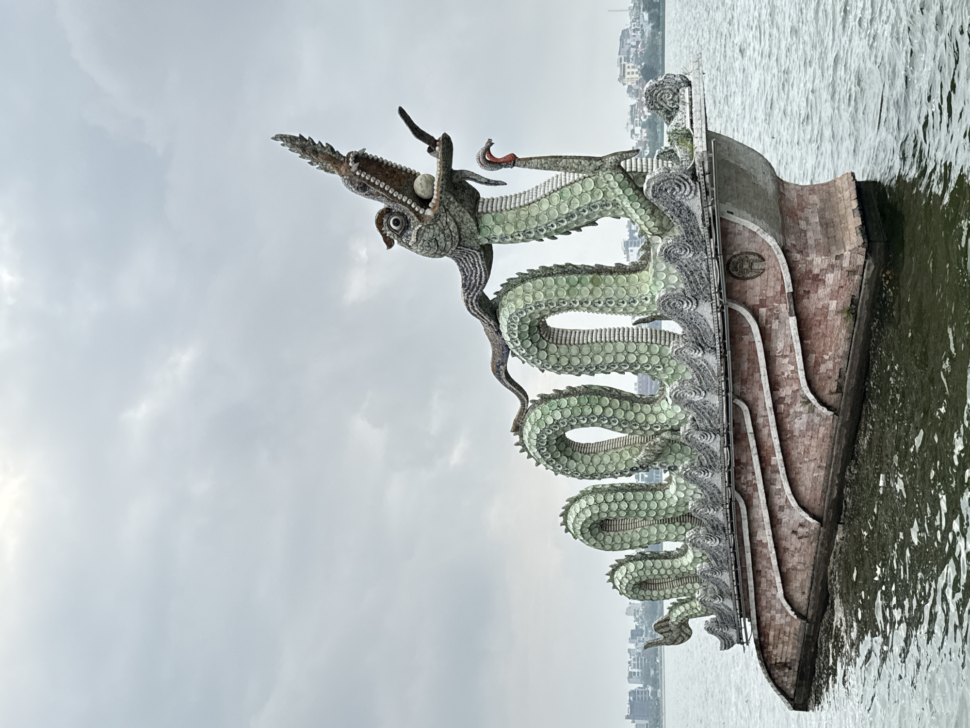 The image shows the dragon boat on West Lake in Hà Nội, Vietnam. The sculpture features a green dragon coiled on the roof of a traditional boat. The water around the boat is rippled, and the sky is overcast. Buildings are visible on the distant shore.
