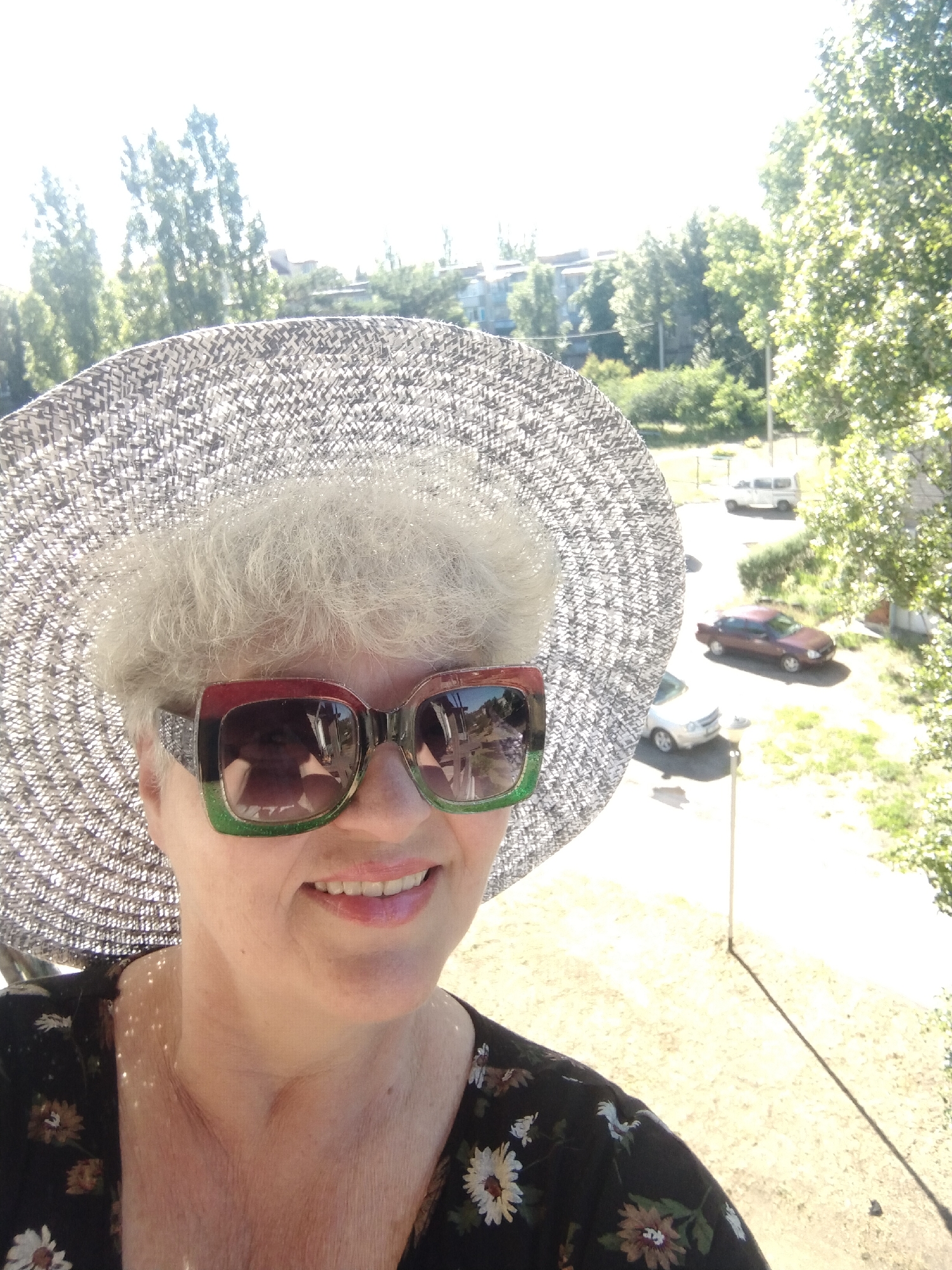A woman is taking a selfie outdoors in Horishni Plavni, Ukraine. She is wearing a wide-brimmed hat and large, colorful sunglasses. She is wearing a dark, floral-patterned top. In the background are trees and buildings, a white van, a red car and a white car can be seen in the parking lot. The scene is bright and sunny.
