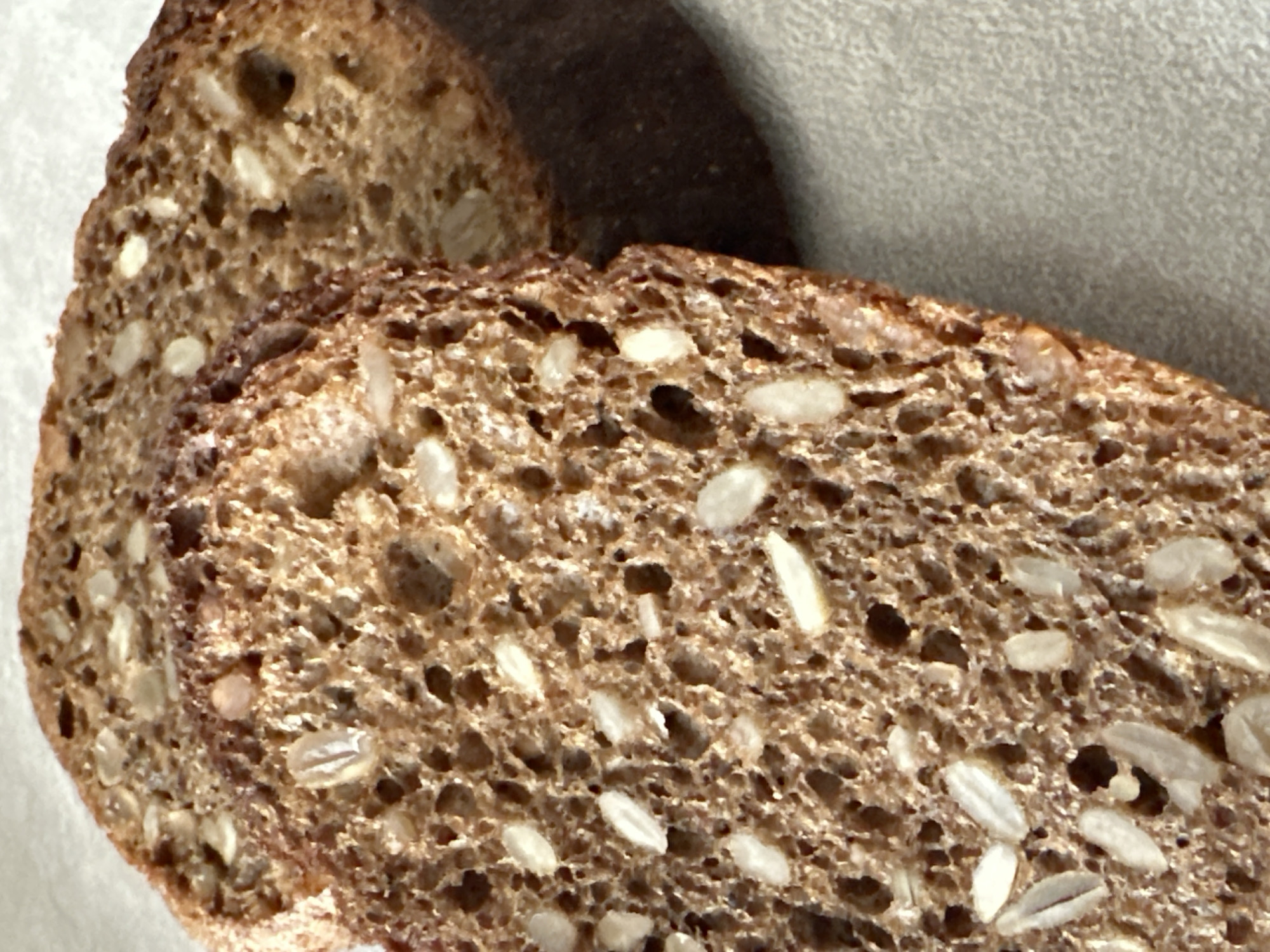 The image shows two slices of dark brown, multigrain bread with visible seeds and air pockets. The bread is positioned close to the camera, filling most of the frame. The slices are slightly overlapping, with the rear slice partially obscured by the front one. The background appears to be a solid, light gray or beige surface. There is a dark shadow cast on the background behind the bread. The scene is indoors, under bright lighting. The location is unknown. There are no people, actions, or interactions in the image.