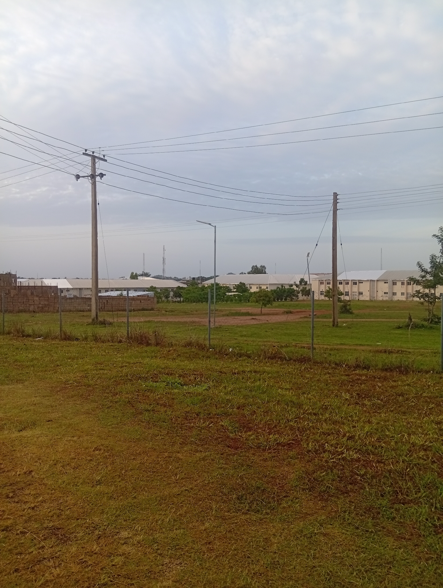 The image depicts an outdoor scene with a cloudy sky. In the foreground, there is a grassy area with patches of dry and green grass. A fence made of metal posts and wires runs across the middle ground. Beyond the fence, there are several utility poles with power lines stretching across the frame. In the background, there are multiple buildings with light-colored walls and roofs. Some trees and vegetation are visible between the buildings and in front of them. A brick wall is partially visible on the left side of the image, separating an area from the open field. The overall environment appears to be a semi-urban or suburban setting with infrastructure for power distribution.