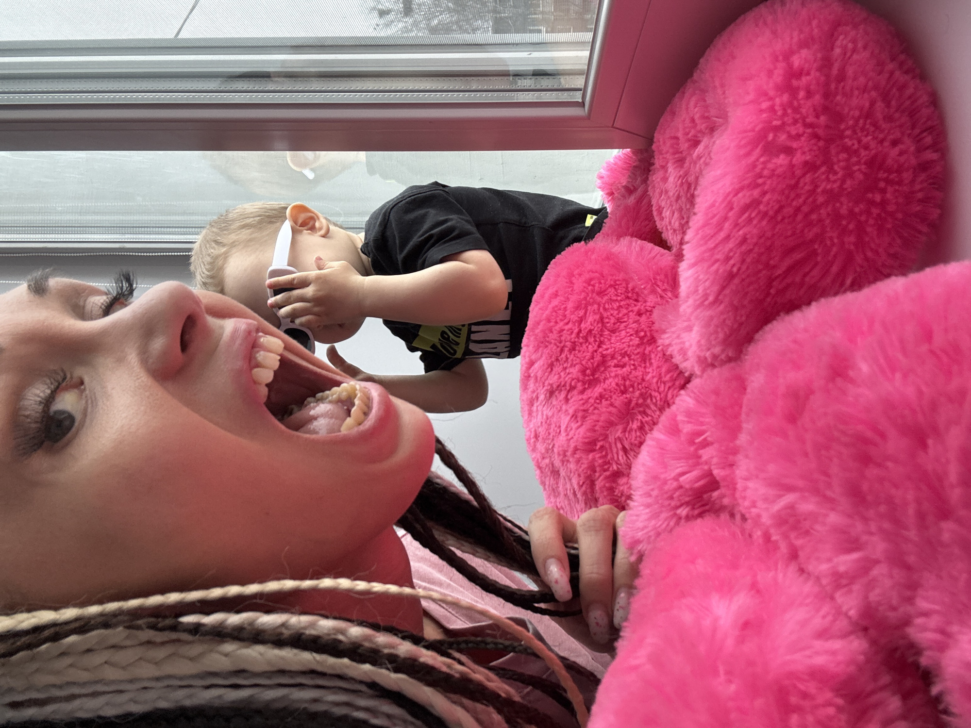 A low-angle photograph shows a woman with multi-toned braided hair lying down, with her mouth open. A child wearing sunglasses and a black t-shirt is seen from the chest up, positioned above her near a window. A large pink, fluffy object, possibly a stuffed animal, is visible next to her. The location is unclear, and there is no definitive evidence indicating it is Rusanovo, Russia.