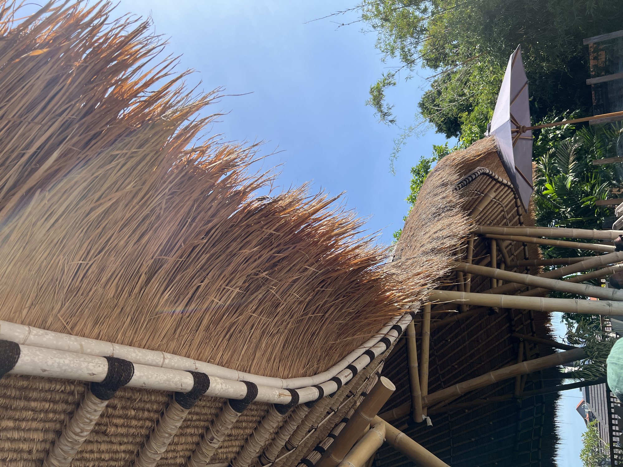 The image shows a section of a traditional thatched roof structure, likely a building or shelter, against a clear blue sky. The roof is constructed with a bamboo frame and covered in dried grasses or straw. Black bindings secure the bamboo elements together. A white parasol is partially visible on the right side, indicating an outdoor setting. Trees and foliage are seen in the background. The location is North Kuta, Indonesia, suggested by the architectural style and tropical vegetation.