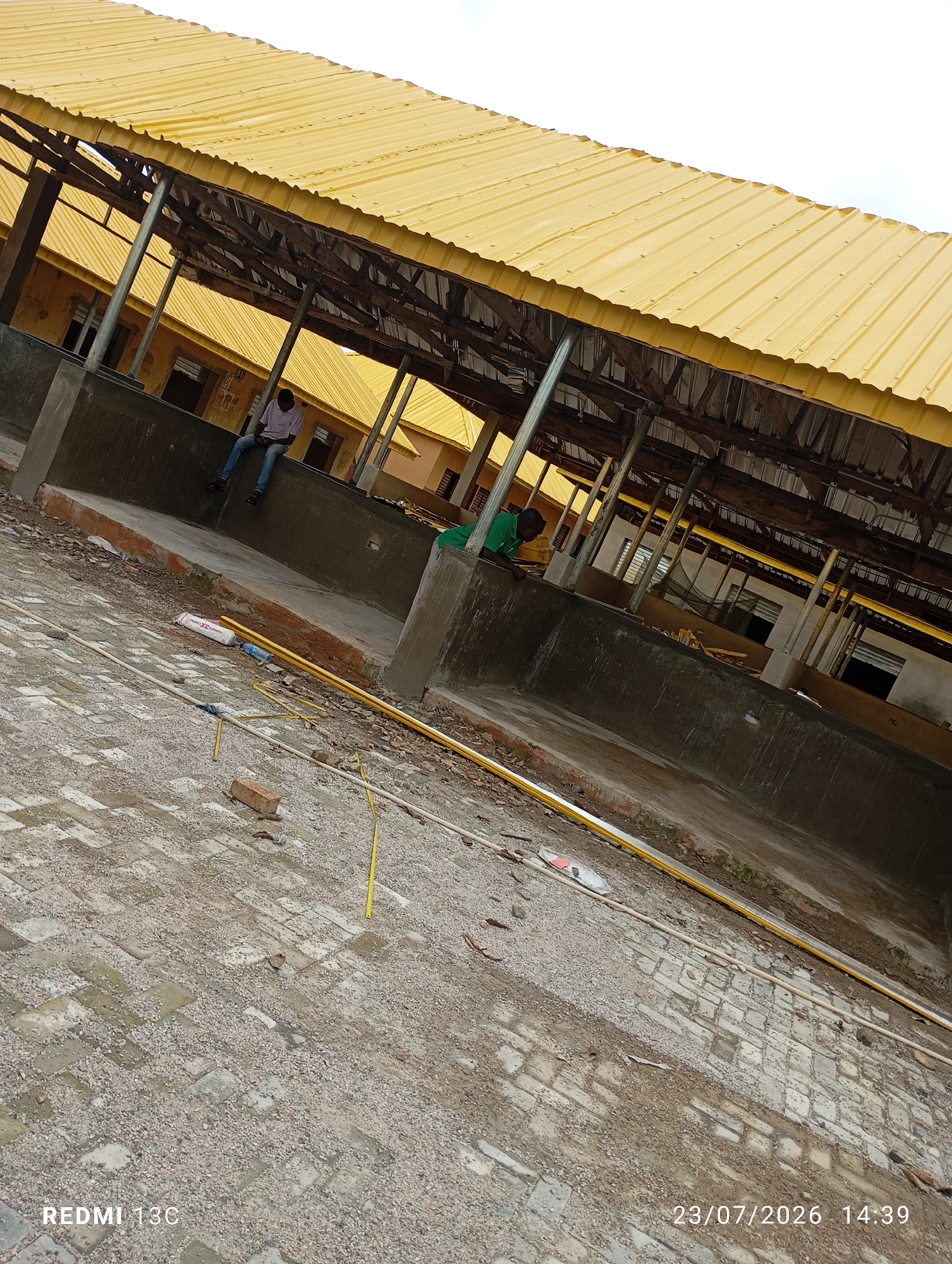 In the city of Ida, Nigeria, a construction site is captured, featuring a partially built structure with a prominent yellow corrugated metal roof supported by metal poles and wooden beams. The ground is covered in paving stones and gravel, indicating ongoing work. Two individuals are present: one man in a light-colored shirt and jeans sits on a concrete ledge, looking down at a handheld device. Another man, wearing a green shirt, leans over a concrete partition, seemingly engaged in work. Various construction materials, including yellow pipes, white tubing, and a brick, are scattered across the site. The overall scene suggests an active construction environment under a cloudy sky.
