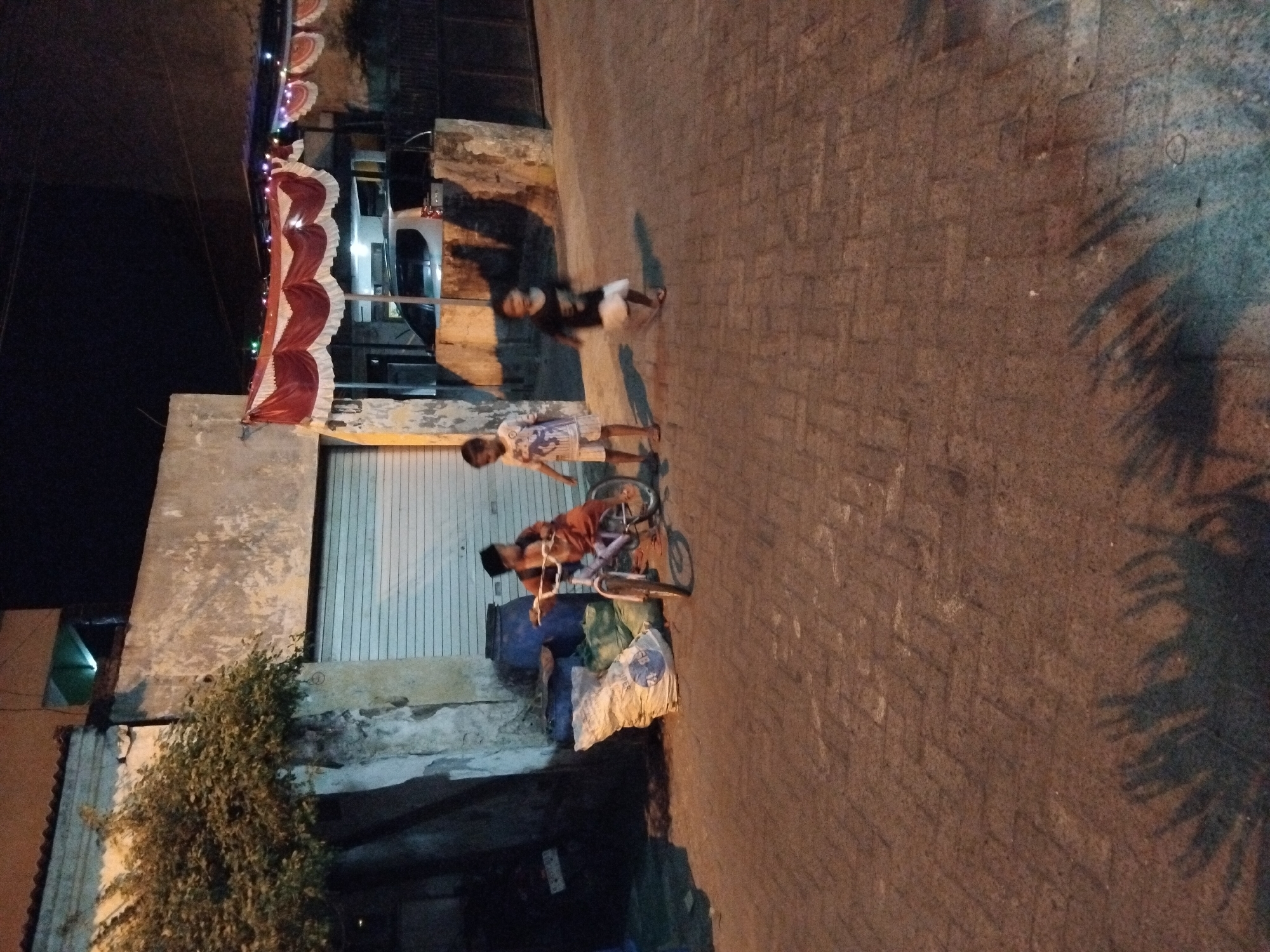 In the city of Kepuh Kiriman, Indonesia, an outdoor scene unfolds at night, illuminated by artificial light. The foreground is a paved walkway with a brick pattern. To the left, a pile of sacks and large blue barrels are visible against the wall of a building. Further to the left, a tree with dense foliage grows next to the building.
The middle ground features three young individuals. A boy on the left, wearing a reddish-brown outfit and a dark cap, is seated on a bicycle. To his right, another boy stands wearing a white t-shirt with a graphic print and white shorts. A third child, to the right of the second boy, is wearing a black t-shirt and white shorts, appearing to be in motion.
The background reveals a building with a large, closed roller door. Above the door, a red and white tented canopy with decorative lights is stretched across the facade. A white vehicle is parked behind the canopy, and a dark fence is visible to the right. The overall ambiance is that of a residential or commercial street at dusk or nighttime.