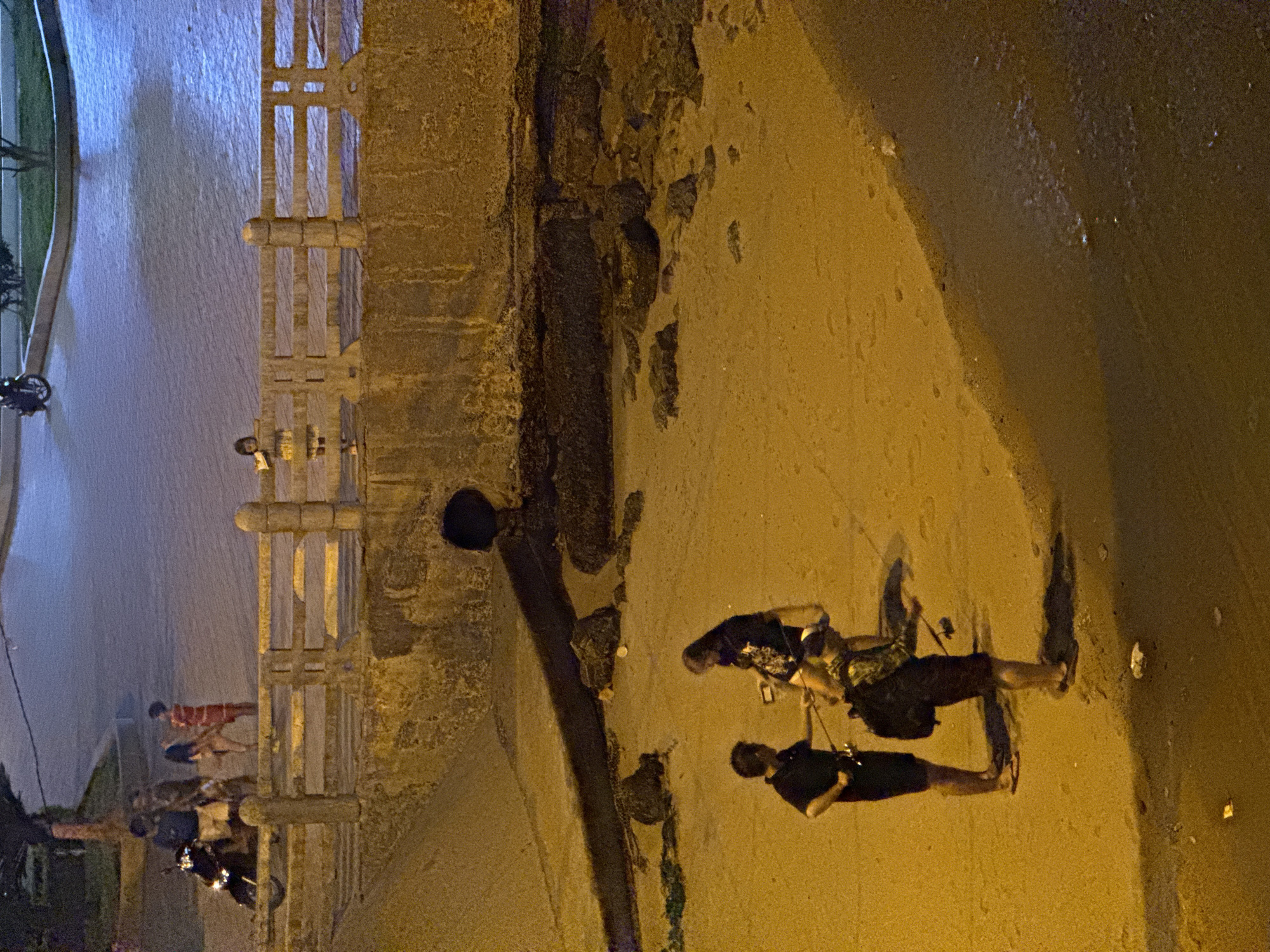 The image depicts a nighttime scene on a sandy beach in Nha Trang, Vietnam, likely near a promenade or elevated walkway. In the foreground, three individuals are visible on the sand, appearing to be engaged in fishing. One person, wearing a dark t-shirt and shorts, holds a fishing rod. Another person, in a patterned shirt and shorts, is also holding a fishing rod and has a backpack. A third individual stands between them, also appearing to hold a fishing rod.
Further back, on the elevated walkway, several people are present. A group of individuals, including one in a red and white striped outfit, stand near a motorcycle. A child is leaning over the railing, looking down at the beach. The walkway is constructed of concrete and features a railing. The background shows a body of water reflecting the artificial lights. The overall lighting is dim, with artificial illumination casting long shadows. The environment suggests a public area with both pedestrian and vehicular access.