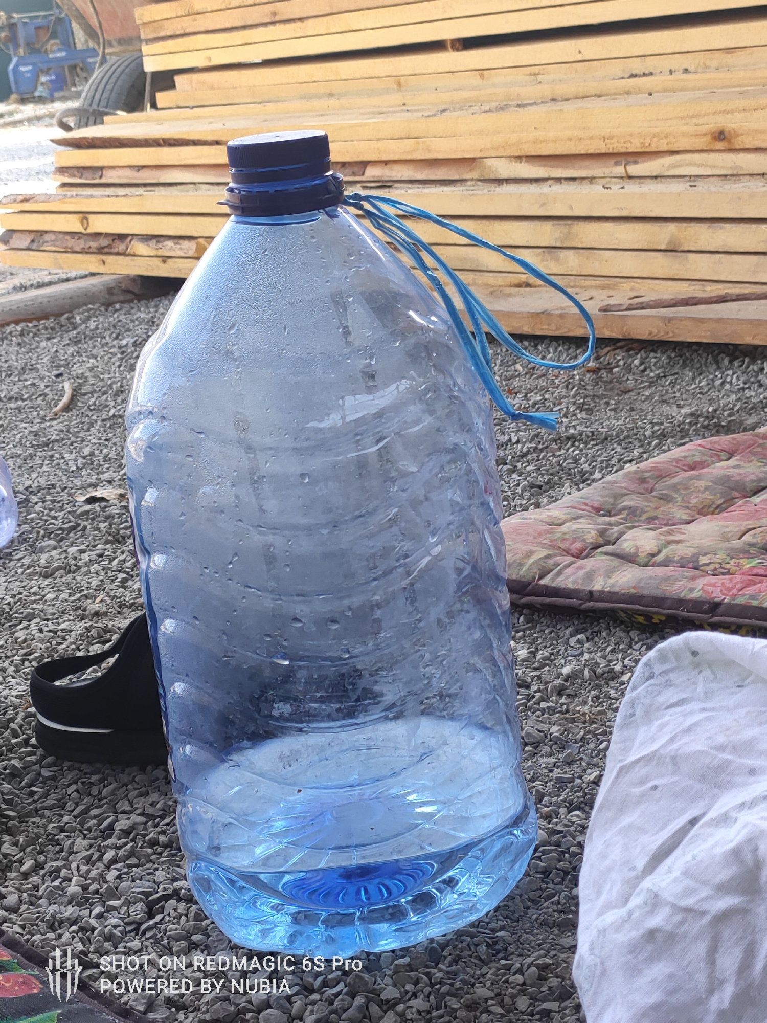 A large, partially filled blue plastic water jug with condensation on its surface sits on a bed of gravel. The jug has a blue cap and a blue string tied around its neck. In the background, a stack of rough-hewn wooden planks is visible, and behind that, part of a blue agricultural machine with a tire. To the right of the jug, a floral patterned mattress or mat lies on the gravel. A black strap, possibly from a bag or shoe, is visible to the left of the jug. A white cloth is on the ground to the right of the jug. The scene appears to be outdoors.