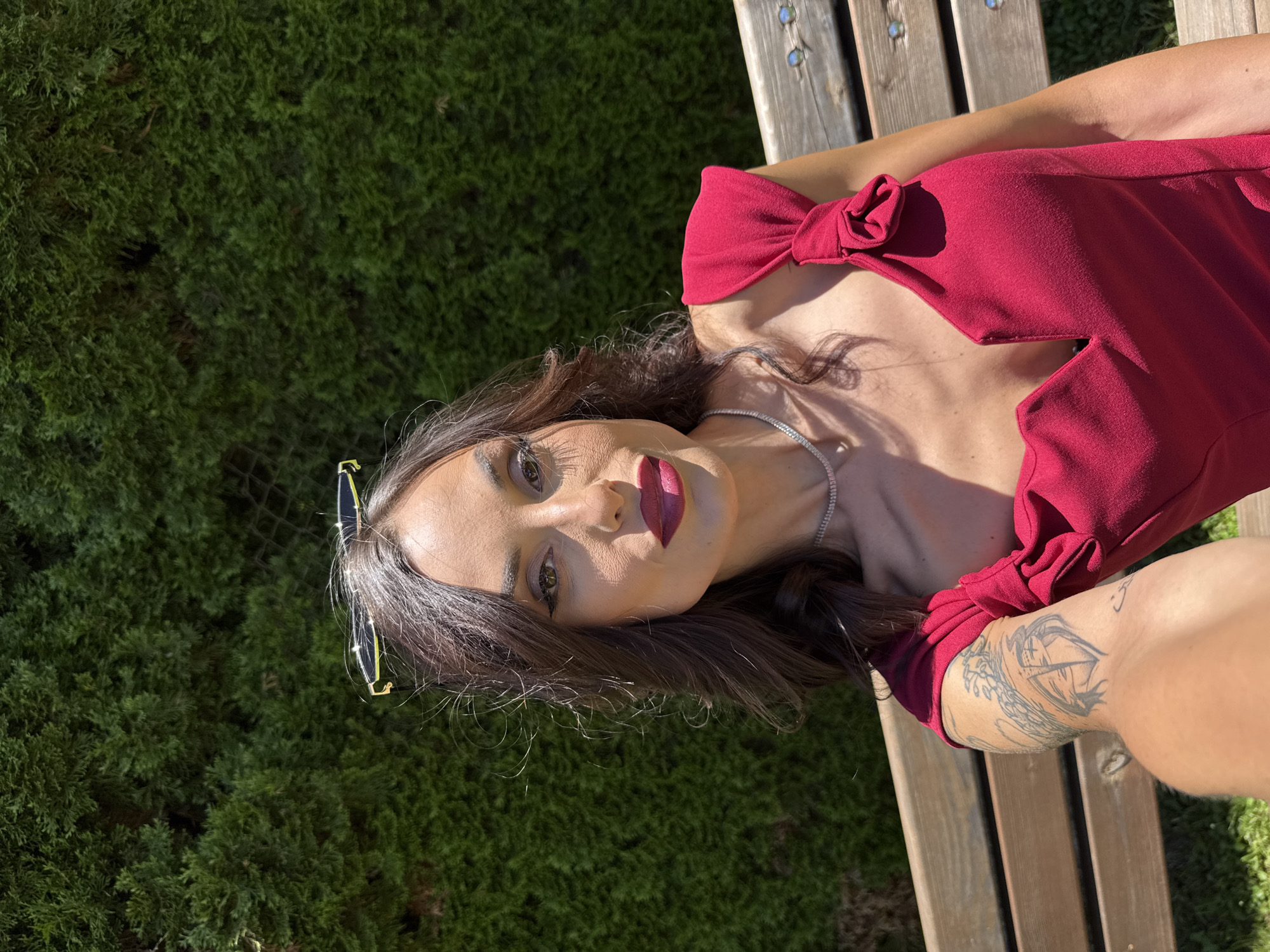 A woman in a red dress reclines on a wooden bench outdoors, with a background of dense green foliage. She is fair-skinned with dark hair and wears sunglasses perched on her head, along with a necklace. A tattoo is visible on her right arm. The scene appears to be captured in daylight with strong sunlight casting shadows. The location is Ribaritsa, Bulgaria.