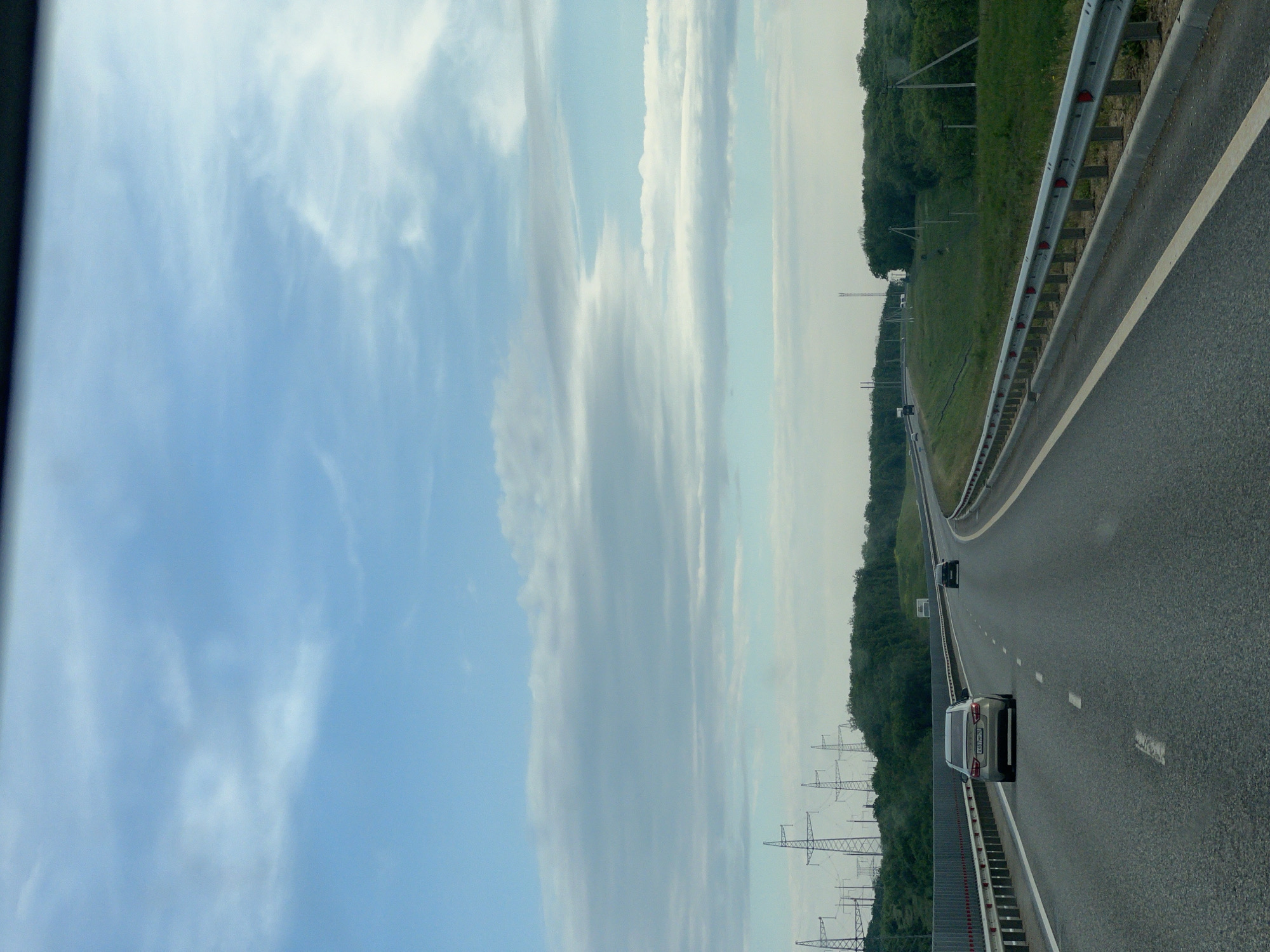 The image shows a multi-lane highway with guardrails running alongside, potentially in the Inkerman, Ukraine area, based on geographical information. Several cars are visible driving on the highway. On the far side of the highway, are a line of electrical pylons, followed by a line of trees. The sky is predominantly blue with scattered clouds. The image appears to have been taken from a moving vehicle.