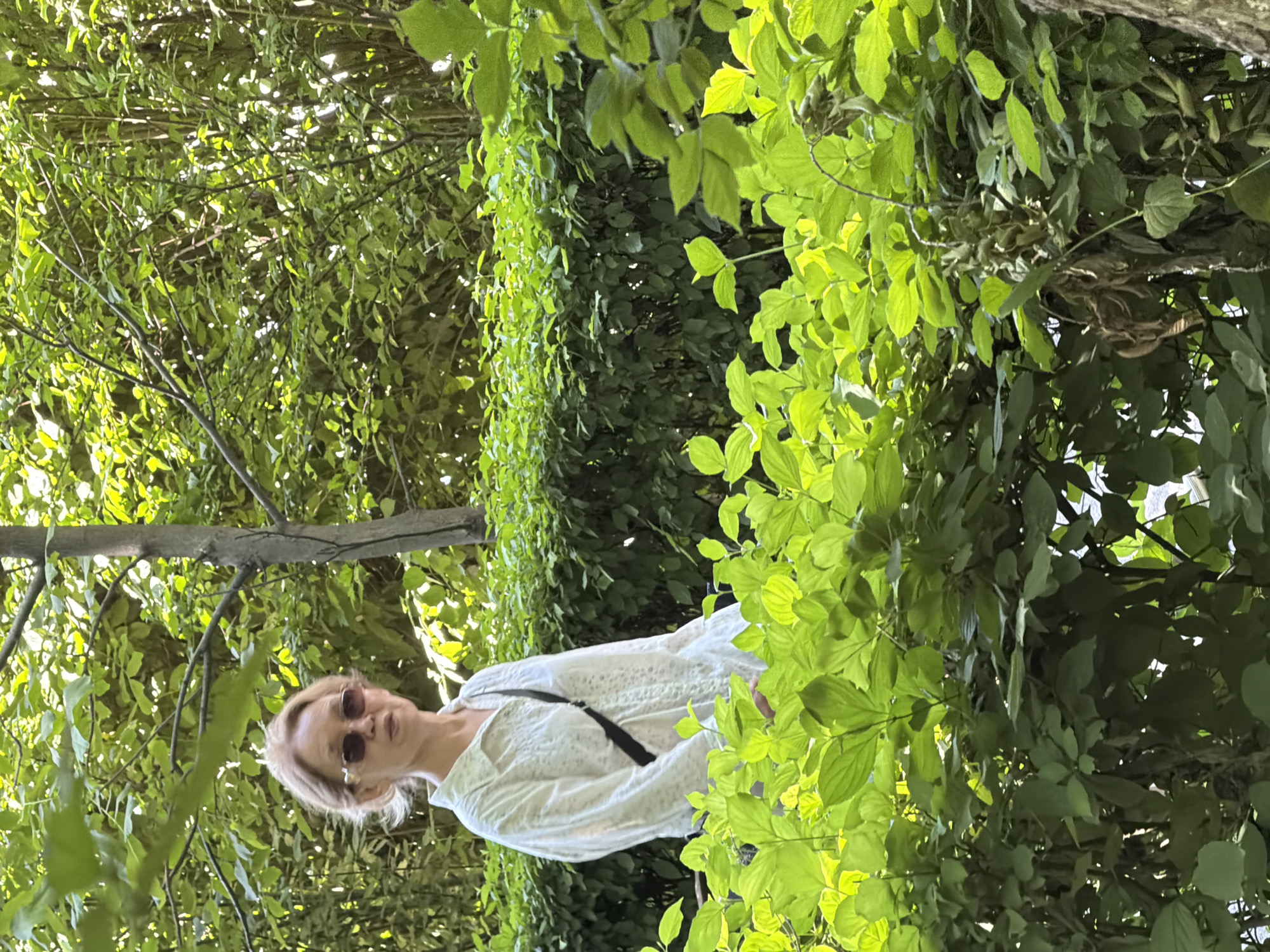 A medium shot captures a fair-skinned woman with short, blonde hair and sunglasses from the chest up. She is wearing a long-sleeved white button-up shirt with a subtle pattern and a black strap across her chest. The woman stands behind a dense, bright green hedge, with a background of more trees and foliage. The lighting suggests it is daytime, with sunlight filtering through the leaves, creating a dappled effect. The setting appears to be an outdoor garden or park within the city.