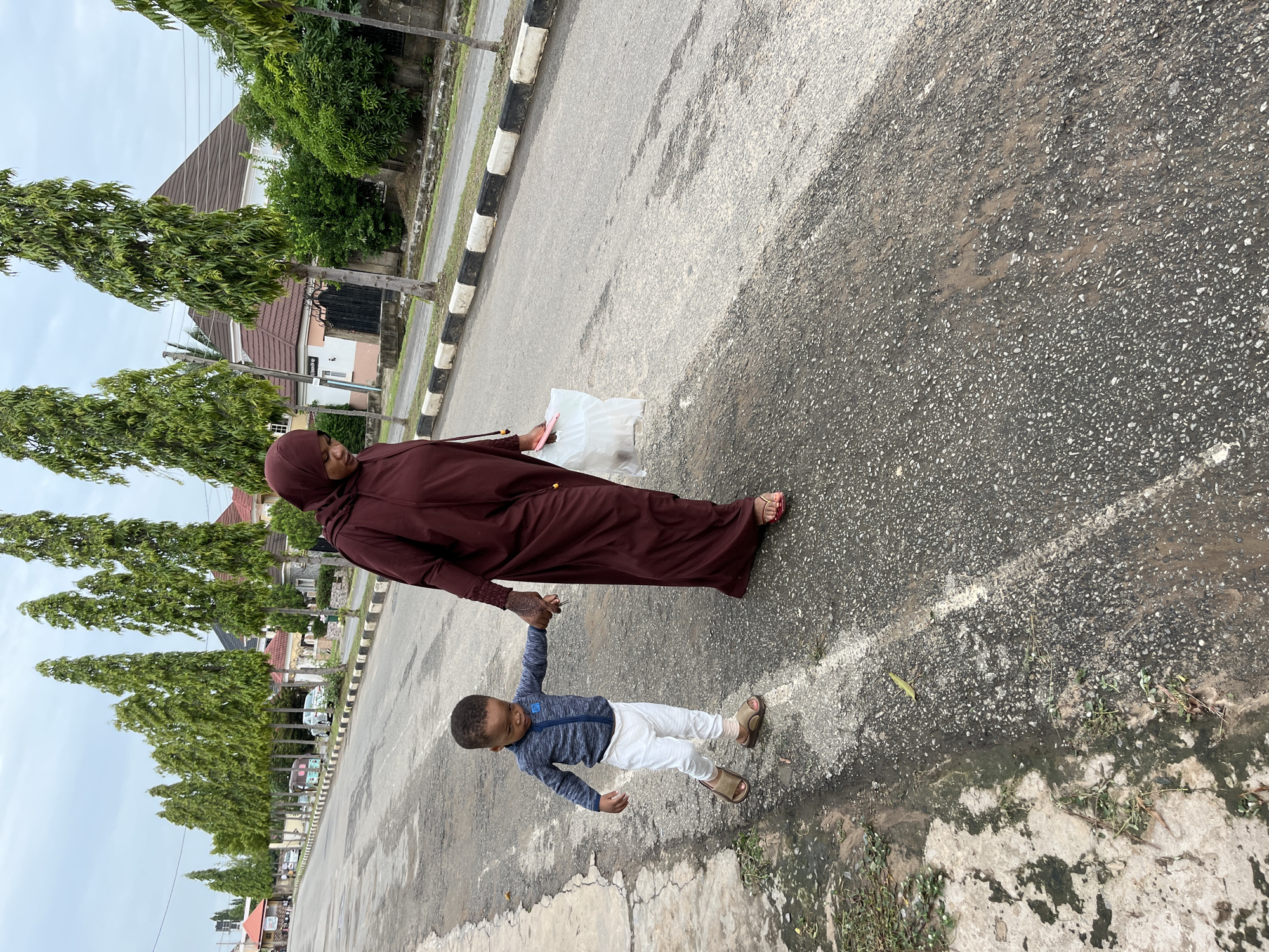 A woman in a maroon hijab and long dress holds the hand of a young boy in a blue shirt and white pants. They are walking on an asphalt road with a black and white curb, bordered by tall, slender trees. The background includes houses and buildings. Based on the urban features and tree type, it is likely a residential street in Abuja, Nigeria. The woman carries a white plastic bag. The ground is wet in places.