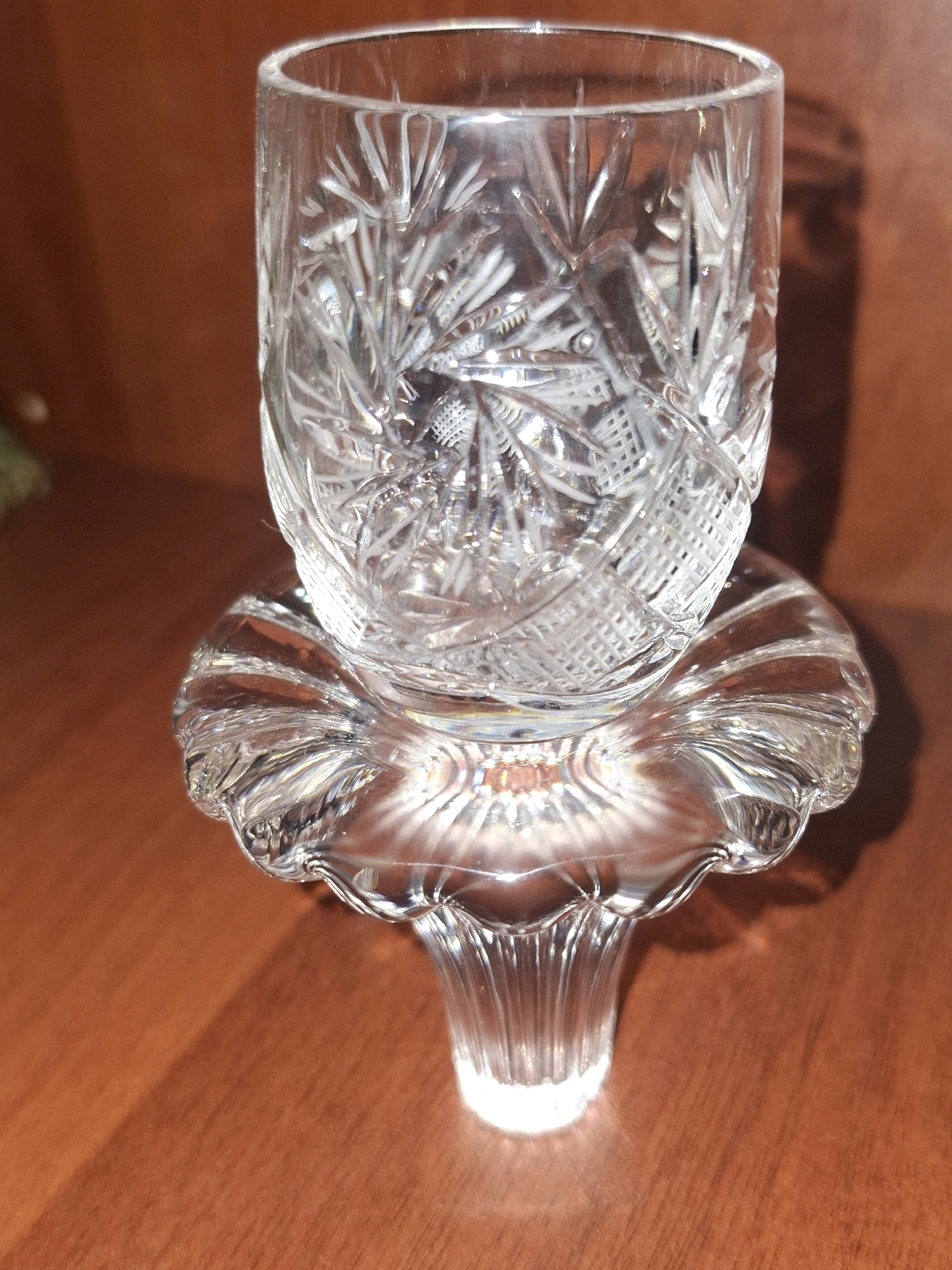 A crystal glass, intricately patterned with starburst designs, rests on a decorative crystal base. The base features a scalloped edge and a fluted stem. Both the glass and its base are clear and highly reflective, capturing and refracting light. The objects are positioned on a polished wooden surface, likely a table or shelf, which has a warm, reddish-brown hue. A shadow of the glass and base is cast onto the surface behind them, indicating a light source from the left. The background is a blurry, out-of-focus brown surface, possibly a wall or another piece of furniture. The scene is indoors and appears to be a still life arrangement. The image was taken in Almaty, Kazakhstan.