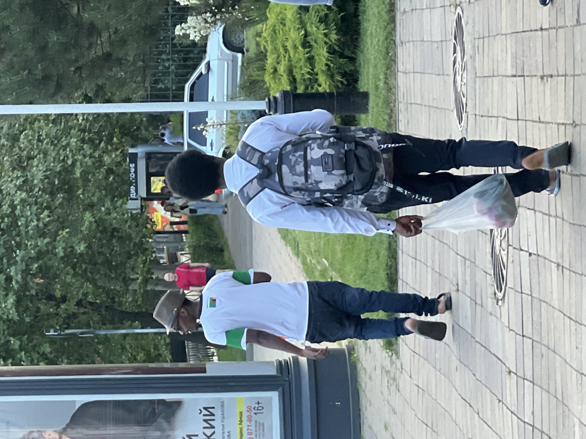 The image captures a street scene in Krasnodar, Russia, likely during daylight hours. In the foreground, two young men are walking away from the camera on a paved sidewalk. The man on the left wears a white t-shirt with green accents on the sleeves and collar, dark blue jeans, and a fedora hat. He is also wearing sunglasses. The man on the right is dressed in a white button-down shirt, black pants, and a camouflage backpack. He is carrying a transparent plastic bag containing what appear to be several items, possibly produce.
In the middle ground, a man in a red t-shirt is walking in the opposite direction. Further back, several other pedestrians are visible near a building with signage that appears to read "ДИН. КОФЕ" (Din. Coffee). A white car is parked on the right side of the frame, partially obscured by lush green bushes and foliage. The background consists of trees and a fence. The overall impression is of a typical urban environment on a pleasant day.
