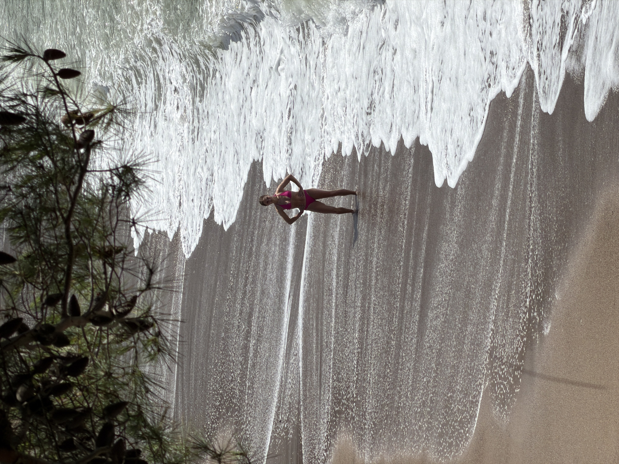 This image depicts a sunny beach scene in Boreti, Montenegro. A woman in a pink two-piece swimsuit stands on the wet sand, positioned confidently with her hands on her hips, facing towards the viewer. She is situated at the waterline where the sea's foamy waves are receding, leaving intricate white patterns on the light-colored sand.
The waves appear energetic and bright, creating a dynamic contrast with the smooth, wet sand. In the foreground, on the left side of the frame, a dark, silhouetted branch of a pine tree, complete with needles and distinct pine cones, partially frames the scene, adding a natural element to the composition.
The strong, bright light suggests it is midday or early afternoon, with ample sunshine illuminating the water and sand. The overall impression is one of a vibrant coastal environment. No text is visible in the image.