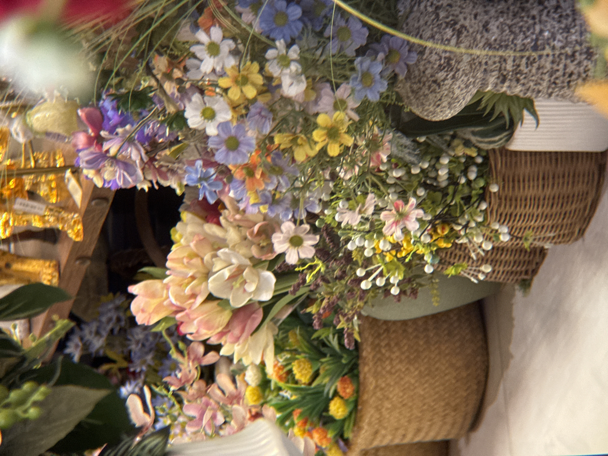 A low-angle, close-up shot captures a display of artificial flowers in a shop. The flowers are arranged in woven baskets and a greenish vase, with a variety of colors including pink, yellow, white, blue, and purple. Some of the flowers are delicate and small, while others are larger and more prominent. The background is blurred, with hints of gold decorative items on a wooden shelf. The floor appears to be tiled with a light-colored pattern. The scene is well-lit, highlighting the vibrant colors of the artificial flora. The location is a flower shop in Nha Trang, Vietnam.