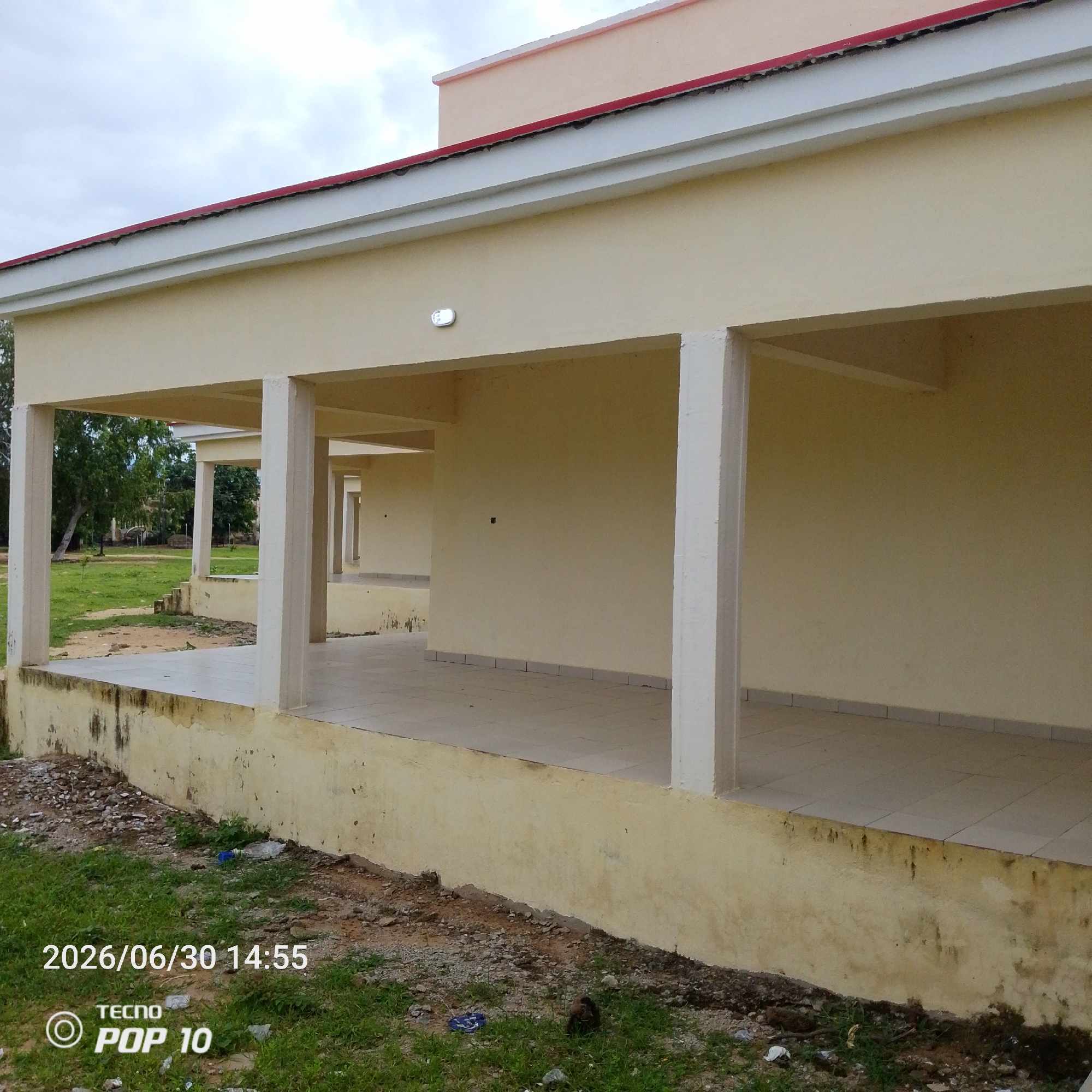 The image shows a covered walkway or verandah of a building with a pale yellow facade. The walkway is supported by regularly spaced white columns and has a tiled floor. Beyond the walkway, there is a grassy area with some trees and vegetation. The sky is overcast, suggesting a cloudy or potentially rainy day. The building appears to be in a state of either recent construction or disrepair, with some visible stains and wear on the lower parts of the walls.
In the foreground, there is a patch of grass and dirt, with some scattered debris, including plastic bottles and other litter. The lower portion of the image contains visible text: "2026/06/30 14:55" indicating the date and time the photo was taken, and a logo with "TECNO POP 10".
The scene is an outdoor environment, likely in a semi-urban or rural setting, given the presence of the building and the natural landscape. There are no people visible in the image. The overall impression is one of a quiet, possibly abandoned or underutilized outdoor space, with a focus on the architectural features of the building and its immediate surroundings. The time of day is mid-afternoon, as indicated by the timestamp.